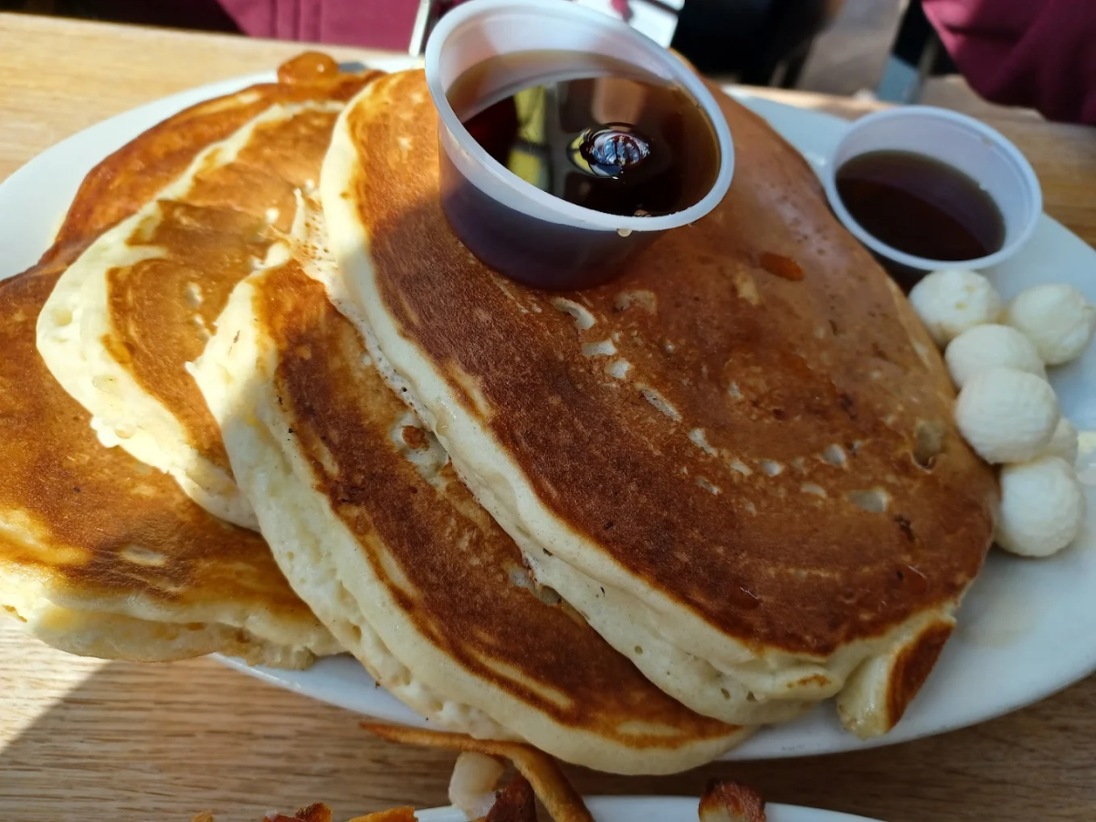 A plate with three large, golden-brown pancakes, a small cup of syrup on top, another cup of syrup on the side, and several small balls of butter on the edge of the plate.