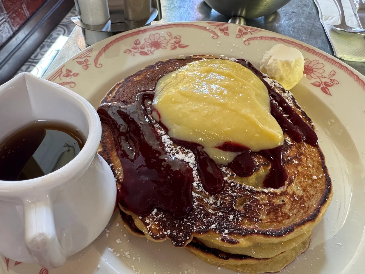 A plate of pancakes topped with yellow custard, red berry sauce, powdered sugar, and a dollop of butter, served with a small pitcher of syrup on the side.