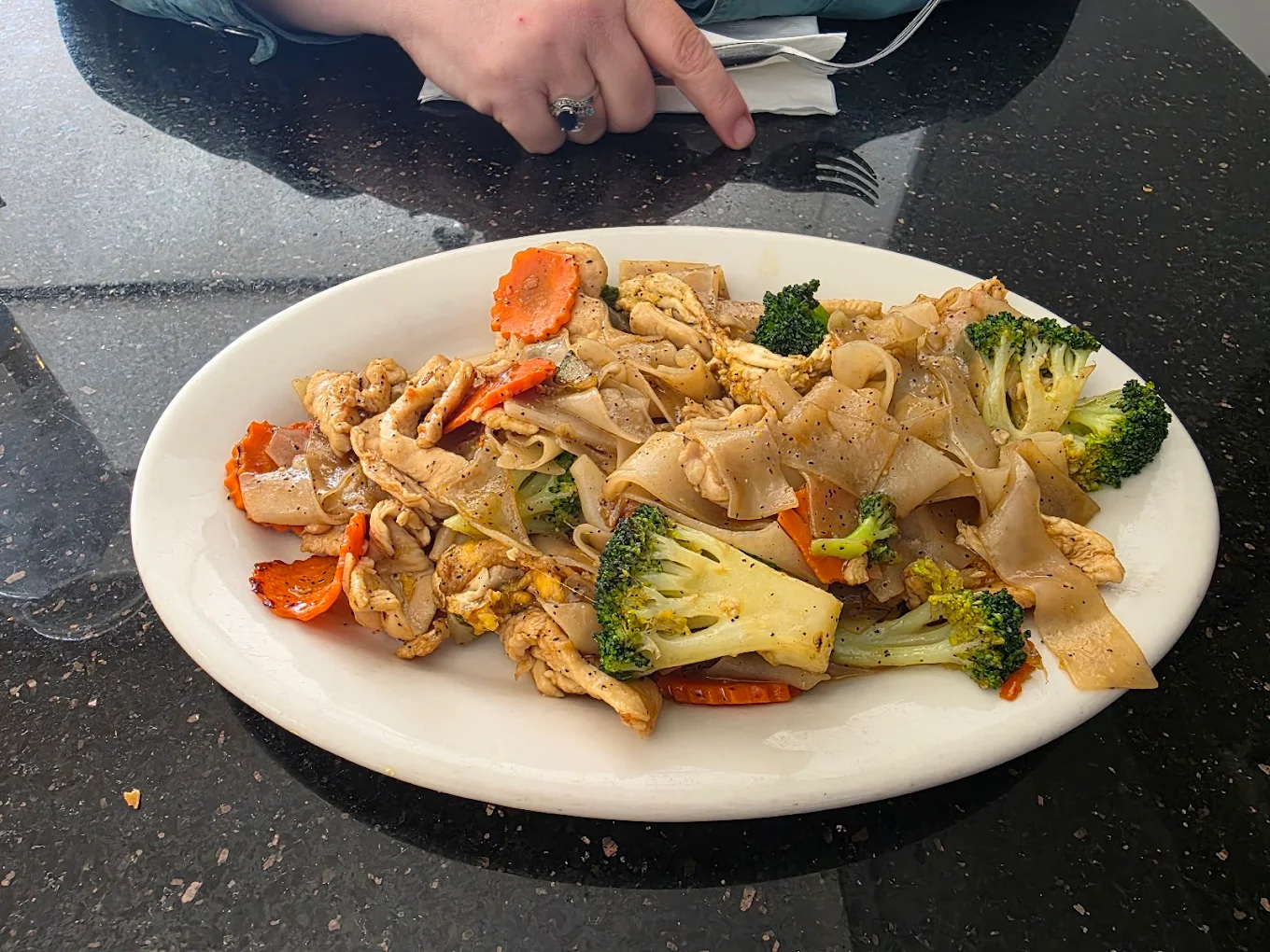 A plate of stir-fried noodles with chicken, broccoli, carrots, and other vegetables sits on a dark countertop. A person’s hand holding a napkin is visible in the background.