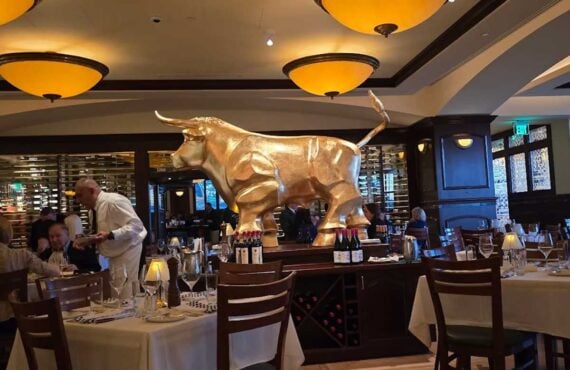 A large golden bull statue, symbolizing strength over bankruptcy, is displayed on a pedestal in the center of an elegant restaurant, surrounded by neatly set dining tables, wine bottles, and warm lighting.