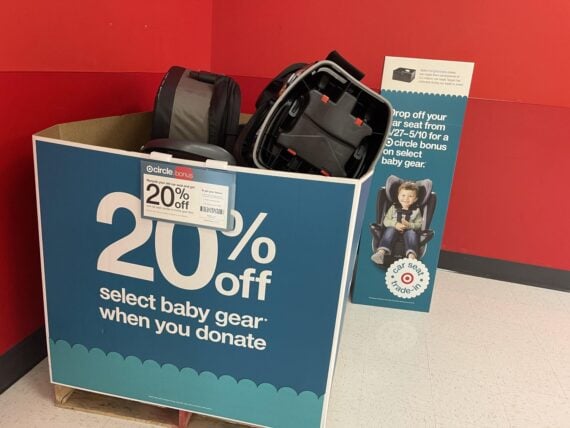 A large blue donation bin labeled &ldquo;20% off select baby gear when you donate&rdquo; contains used car seats. A sign behind the bin displays information about the trade-in offer, all set against a red wall.