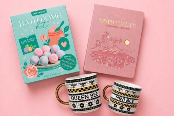 A bath bomb kit, pink mixed puzzles book, and two mugs labeled "QUEEN BEE" and "MINI BEE" with black, white, and yellow patterns&mdash;these charming Aldi Mother's Day gifts are beautifully arranged on a pink background.