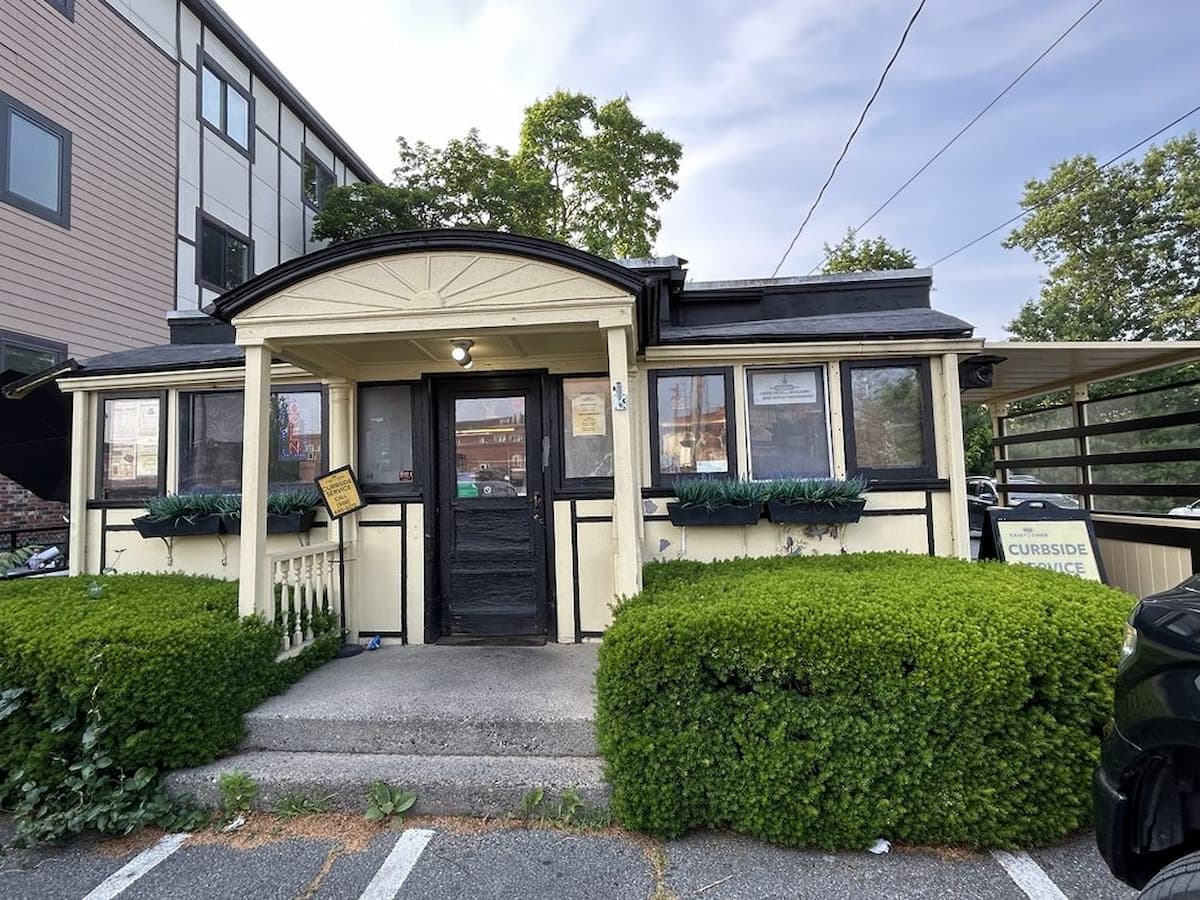 A small, yellow, cottage-style building with black trim and green bushes in front echoes the charm of old-school diners in America. Signs are posted by the entrance, including a "curbside service" sign on the right near the parking lot under a cloudy sky.