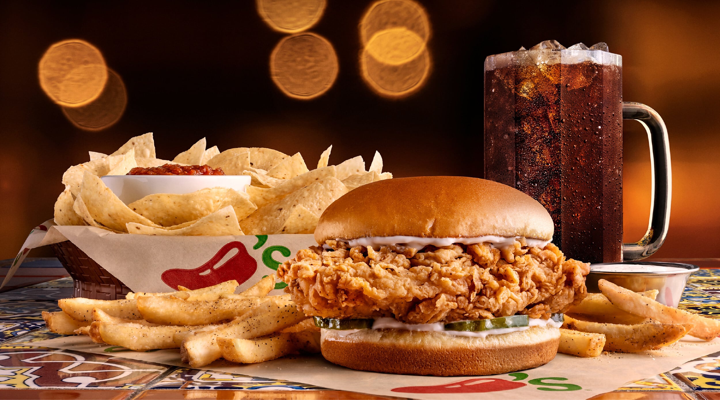A crispy chicken sandwich with pickles and mayonnaise, served with golden fries, nachos with salsa, and a large glass of soda on a decorative tiled table with warm, blurred lights in the background.