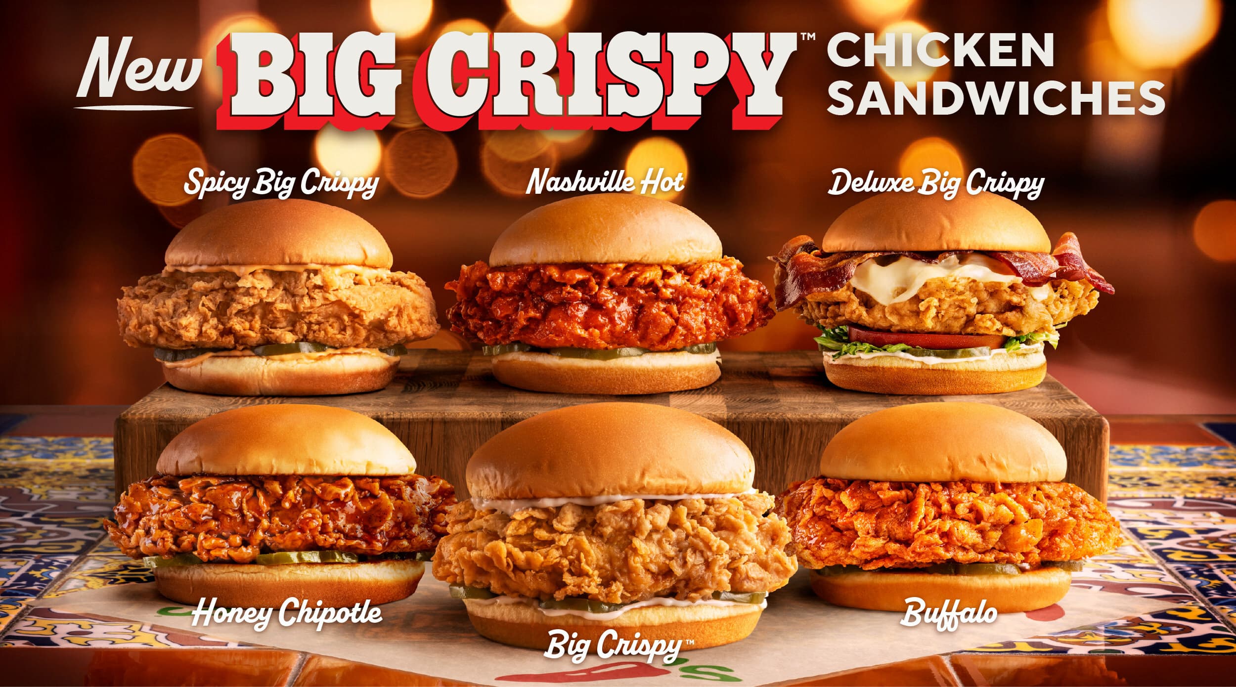 Six chicken sandwiches are displayed on a wooden board and table under warm lights. Each sandwich is labeled: Spicy Big Crispy, Nashville Hot, Deluxe Big Crispy, Honey Chipotle, Big Crispy, and Buffalo.