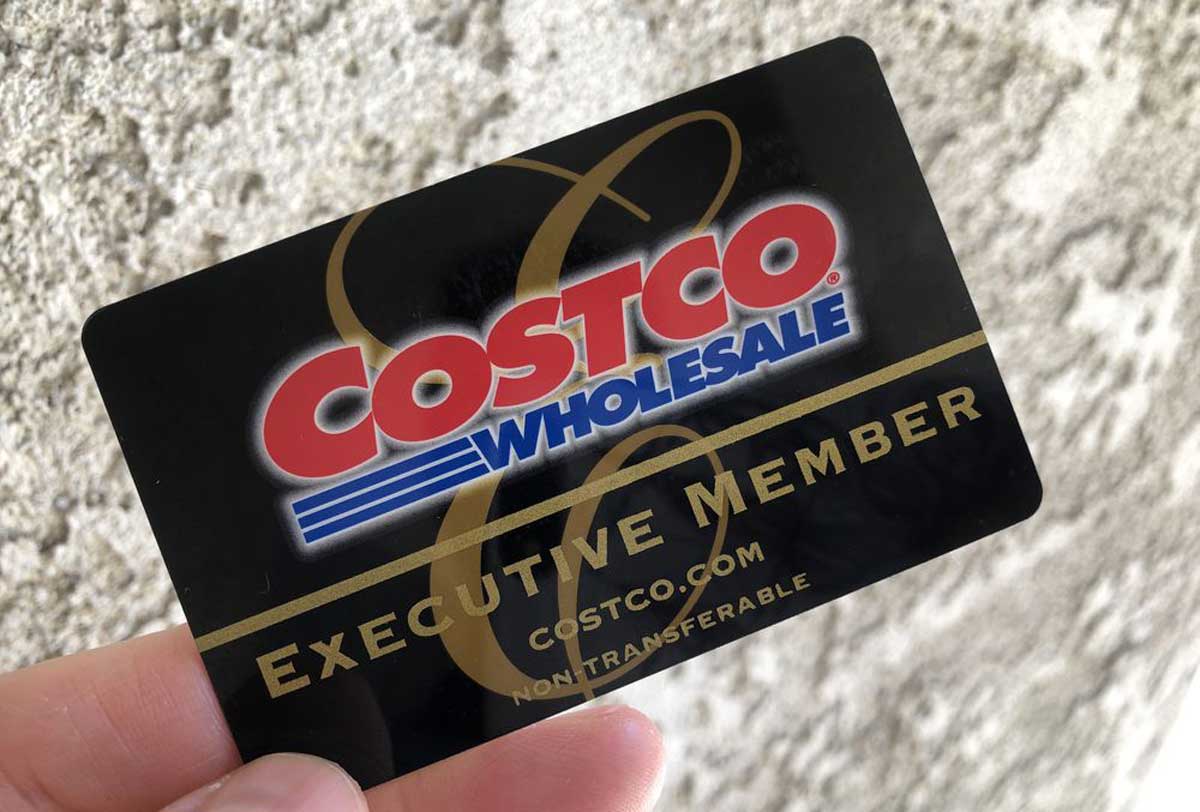 A hand holds a black Costco Wholesale Executive Member card against a textured gray wall background. The card, showcasing gold and red lettering with the Costco logo, offers exclusive costco executive rewards.