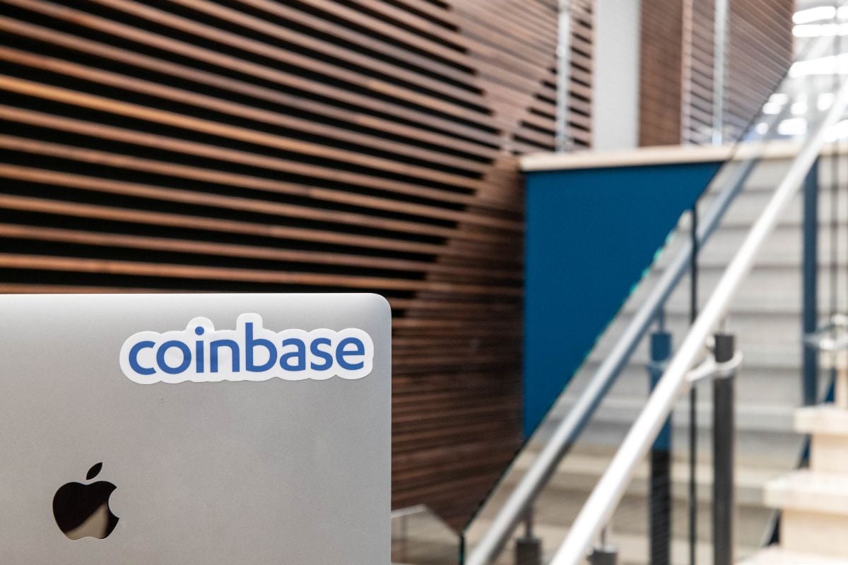 A silver laptop with an Apple logo and a "coinbase" sticker is shown in the foreground, set against modern, wood-paneled walls—subtly reflecting the tension of the 2026 layoffs in the tech workspace beyond.