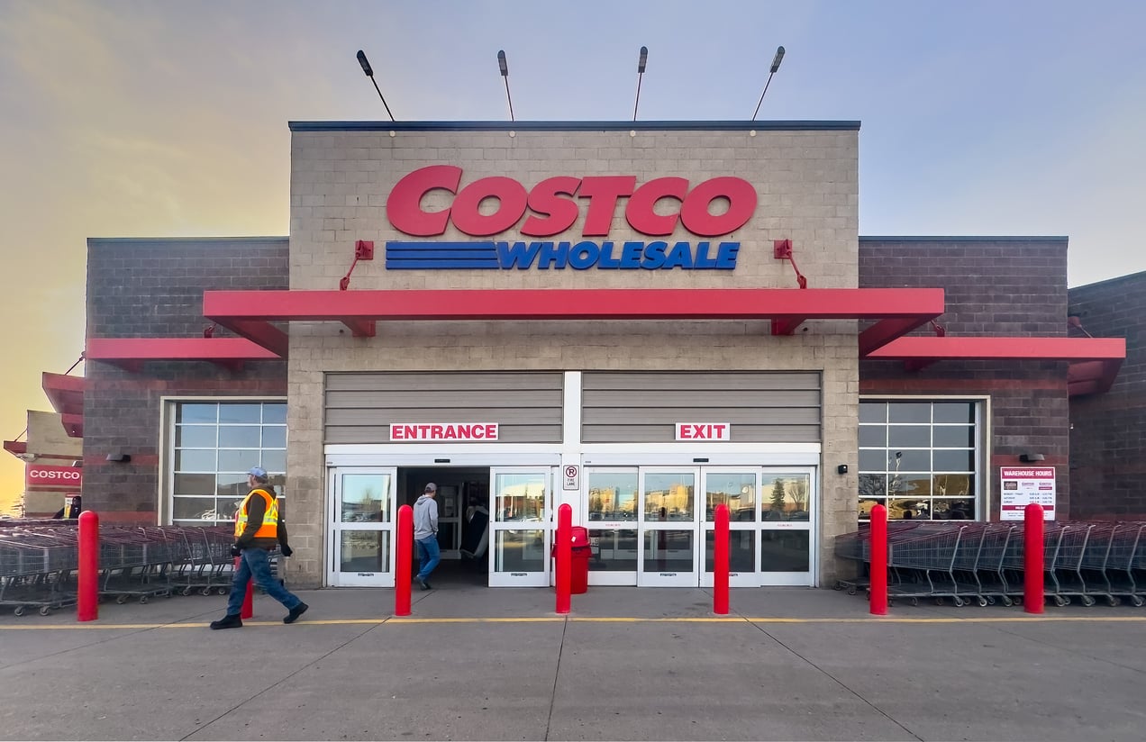 The entrance and exit of a Costco Wholesale store, with shopping carts on the side, red poles in front, and people walking in and out—perhaps pondering popular Costco myths as they shop.