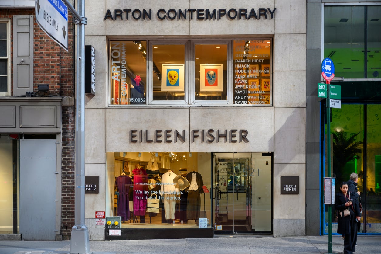 The storefront of EILEEN FISHER clothing store, known among thrift store brands, features window displays above for ART ON CONTEMPORARY gallery with framed colorful artwork. People walk by on the city sidewalk outside.