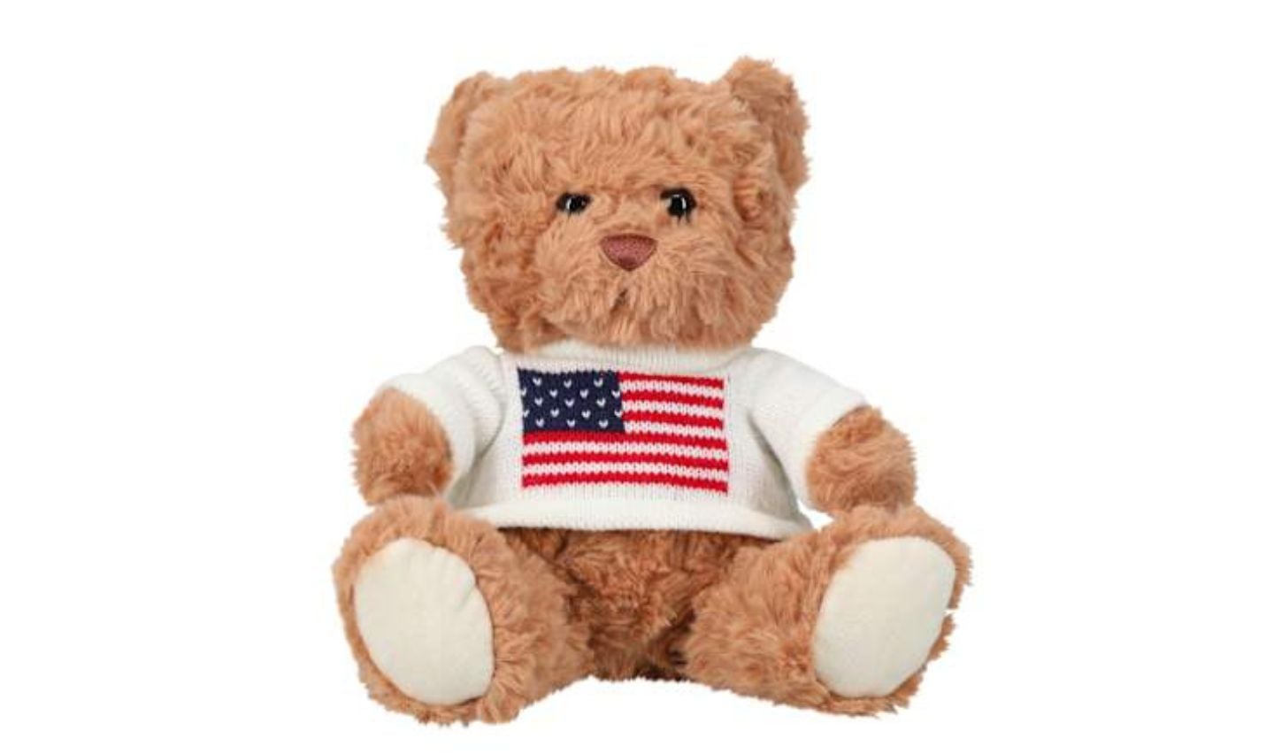 A brown teddy bear wearing a white sweater with an American flag design, part of the new Five Below products, sits against a plain white background.