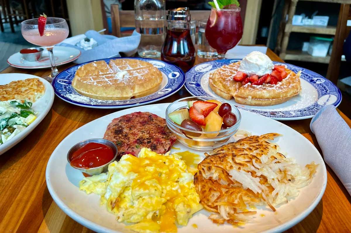 A breakfast spread with scrambled eggs, hash browns, sausage patty, fruit, and ketchup, plus two plates of waffles—one with strawberries and whipped cream, the other plain. Two colorful drinks are also on the wooden table.