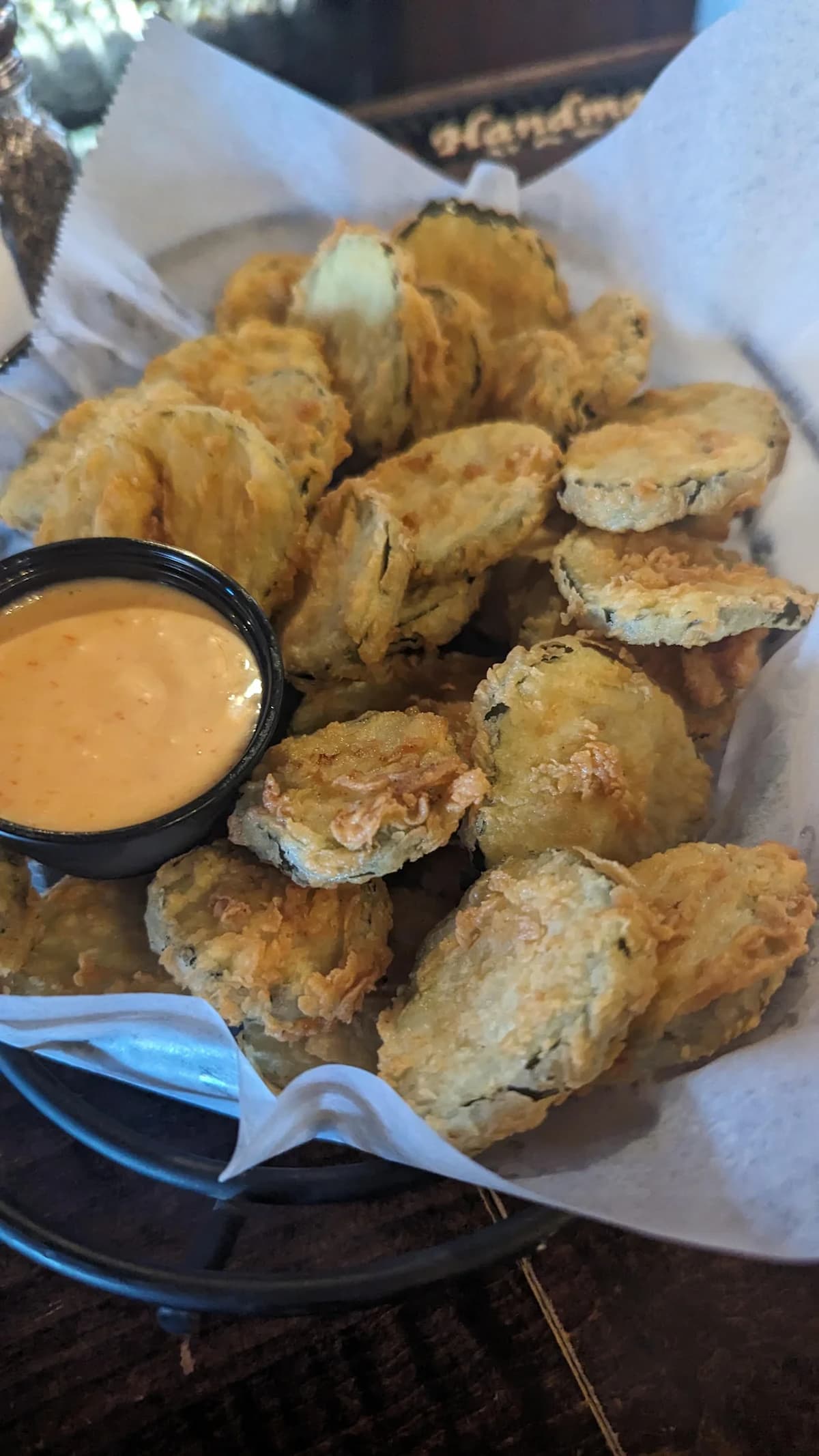 A basket of crispy fried pickle slices served on parchment paper with a small cup of creamy dipping sauce.