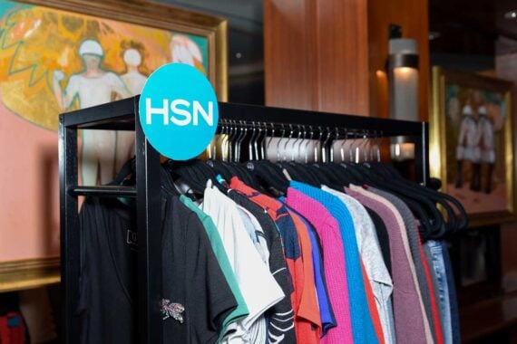 A clothing rack with various colorful shirts on hangers stands beneath a turquoise HSN sign, a vibrant contrast amid uncertainty as whispers of bankruptcy linger. In the background, framed artwork featuring human figures adorns the wall.