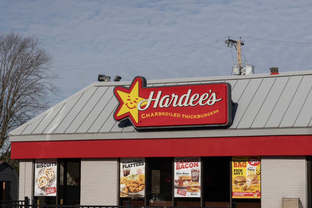 A Hardee’s fast food restaurant with a red and yellow sign reading “Hardee’s Charbroiled Thickburgers” sits under a gray roof. Despite rumors of bankruptcy, menu posters remain visible in the windows below the sign.
