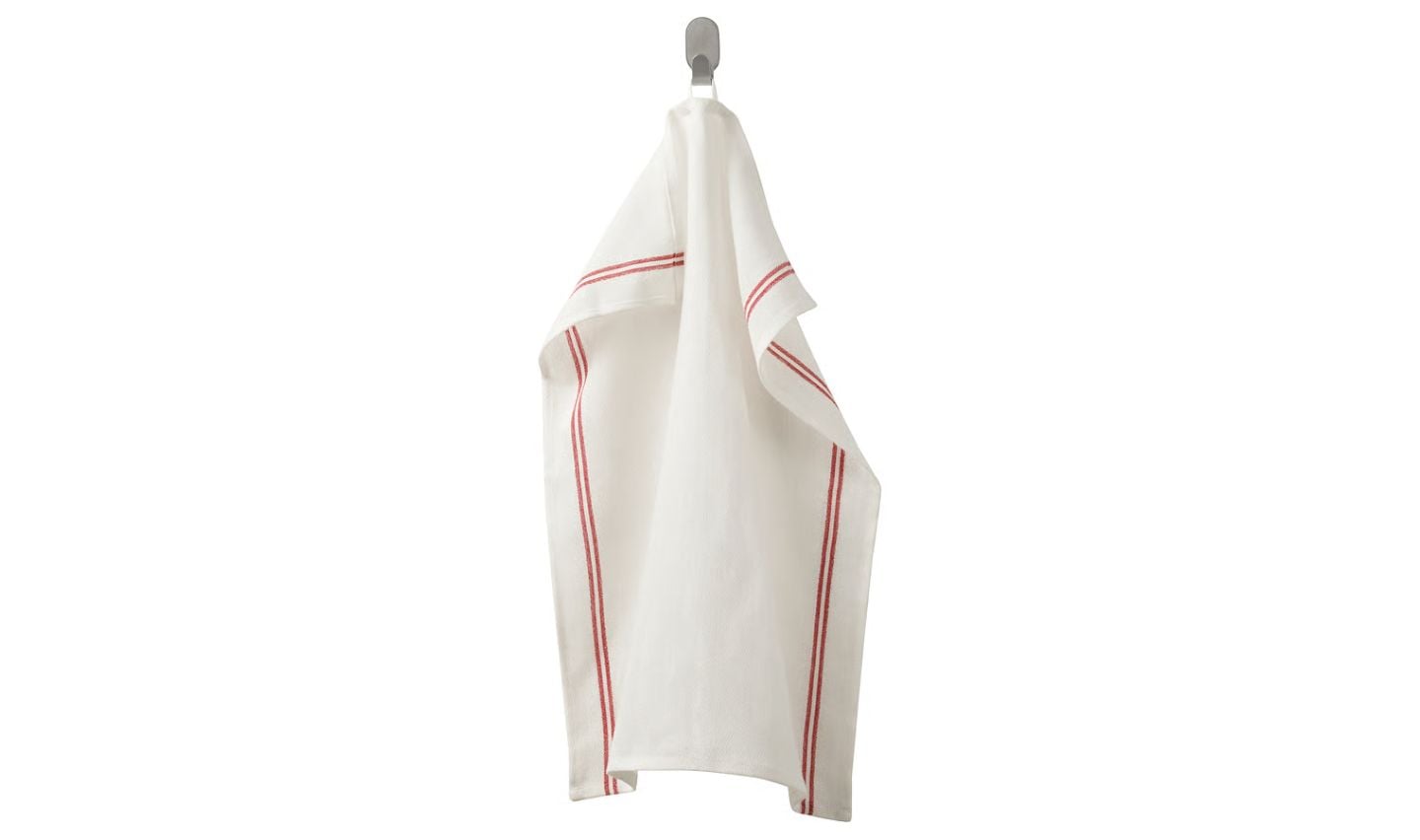 A white kitchen towel with two red stripes near the edges, similar to classic IKEA loss leaders, hangs on a simple wall hook against a plain background.