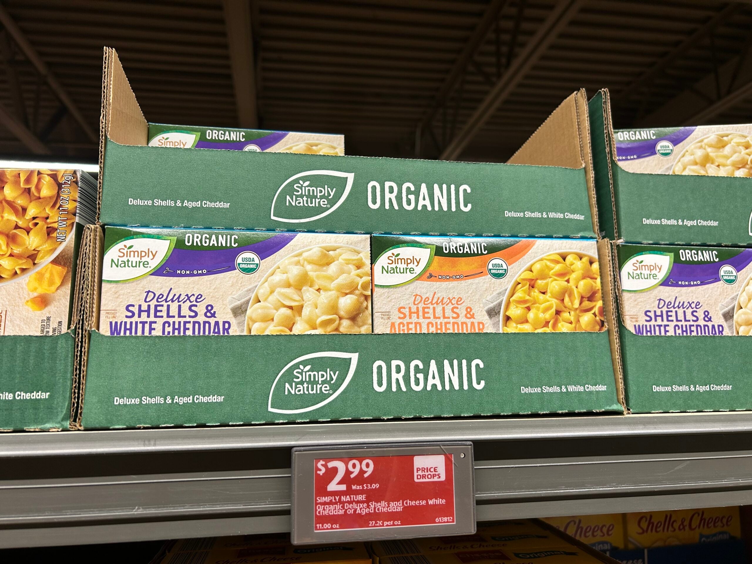 Boxes of Simply Nature Organic Deluxe Shells & White Cheddar and Deluxe Shells & Aged Cheddar are displayed on a grocery store shelf with a price tag of $2.99 below them.