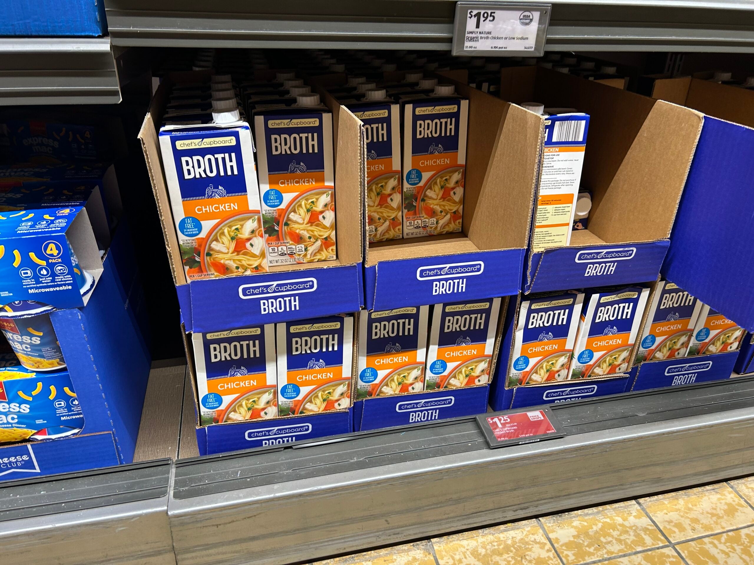 Cartons of chicken broth are stacked on a grocery store shelf, with the label "Broth Chicken" visible. The cartons are mostly in blue and yellow packaging, some in boxes, with a price tag above the shelf.