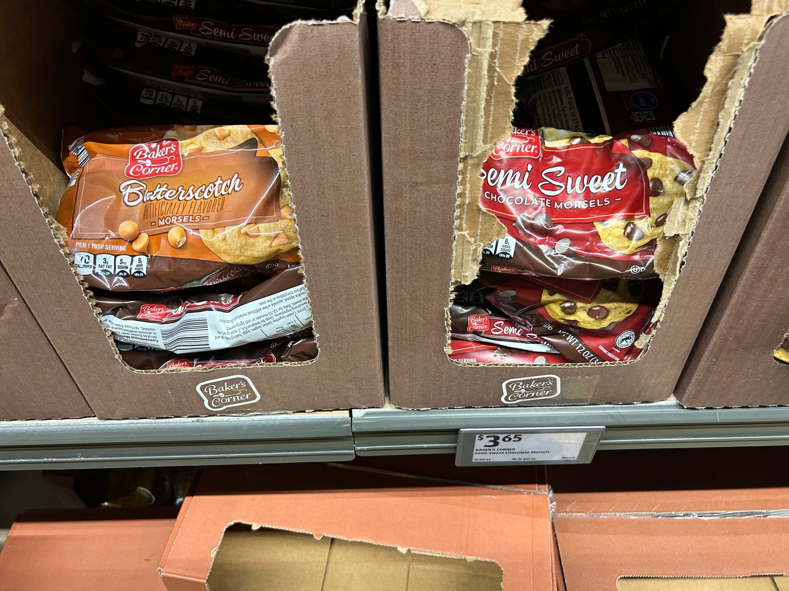 Packages of Baker’s Corner butterscotch and semi-sweet chocolate morsels are displayed in partially opened cardboard boxes on a store shelf. A price tag below shows the cost as $3.65.