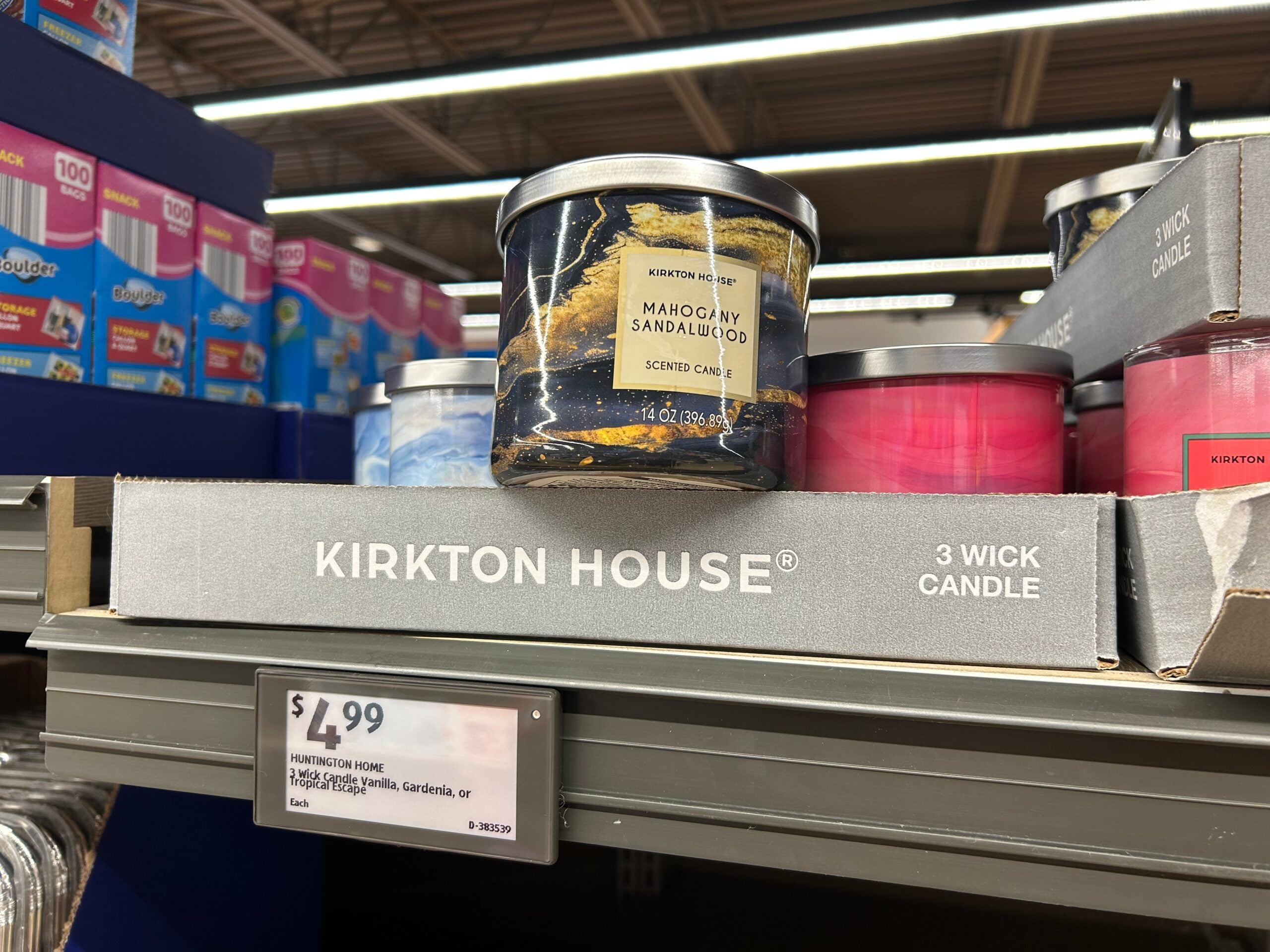 A Kirkton House Mahogany Sandalwood 3-wick candle sits on a store shelf next to other candles. A price tag below shows $4.99. The candle has a marbled black and gold design on the label.