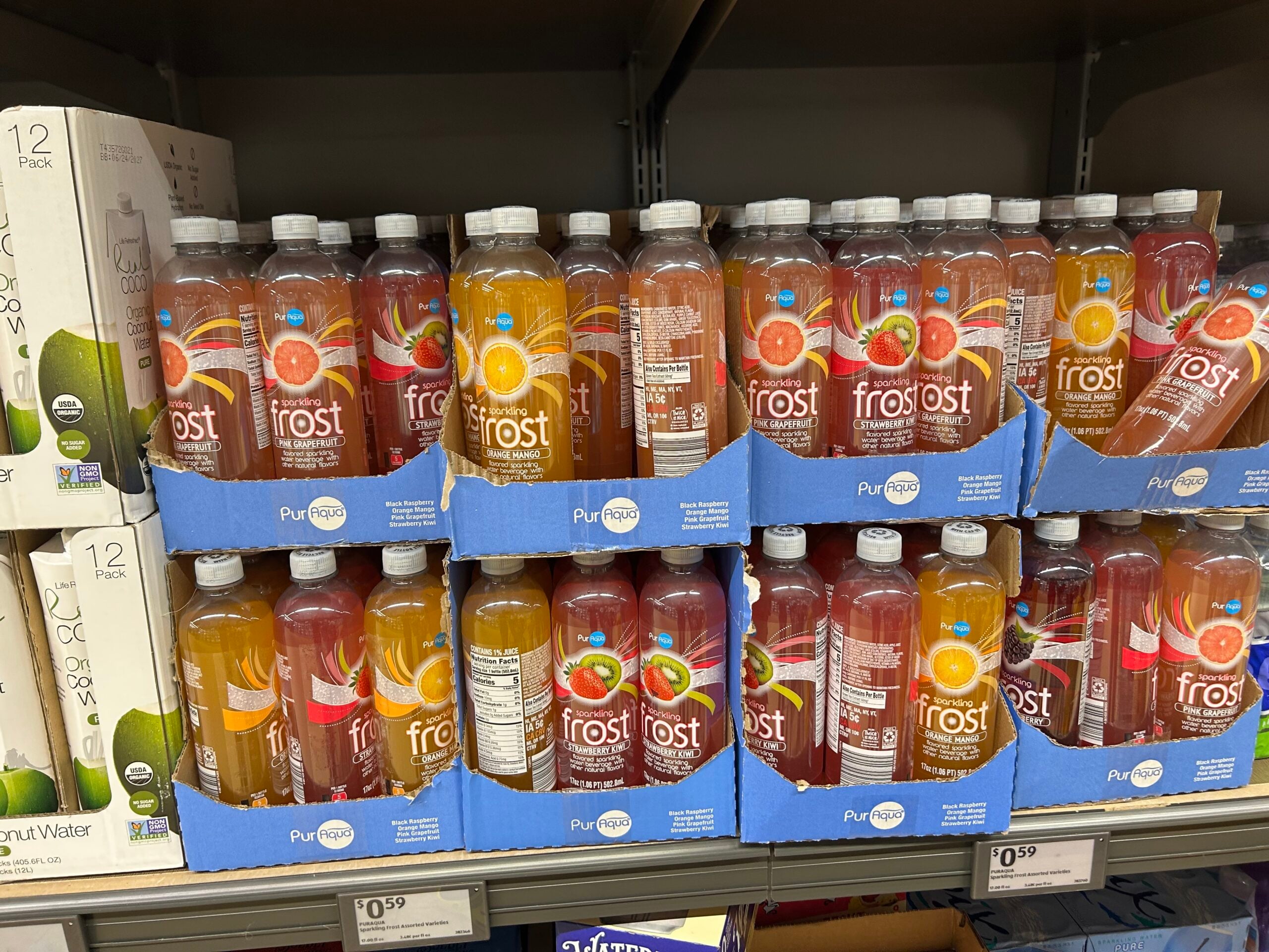 Bottles of Frost flavored sparkling water, mainly in orange and pink varieties, are displayed in blue cardboard trays on a grocery store shelf. The price tag below reads $0.59.