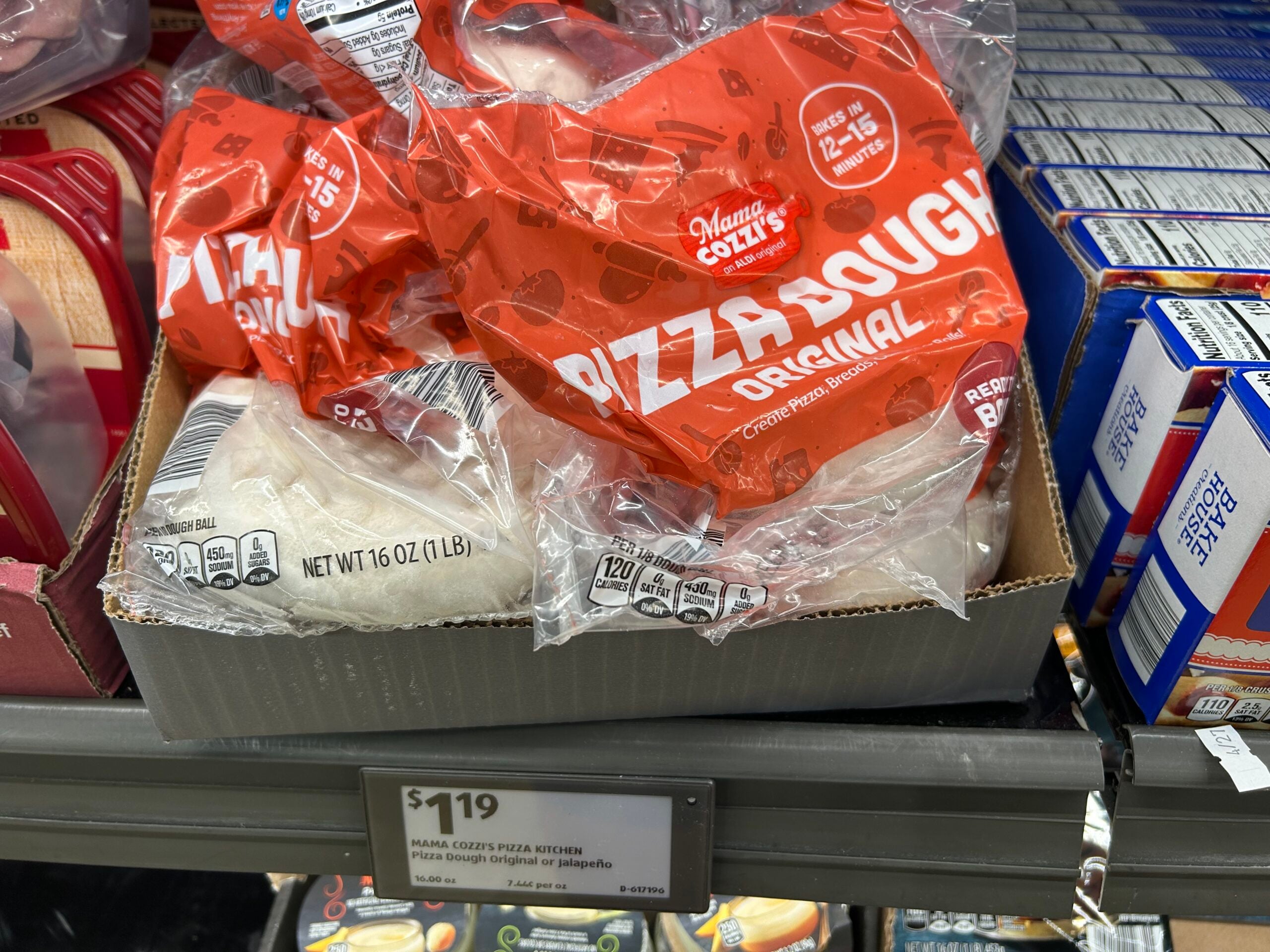 A grocery store shelf displays several bags of Mama Cozzi's Original Pizza Dough in clear and red packaging. The price label below shows $1.19 per 16 oz package.