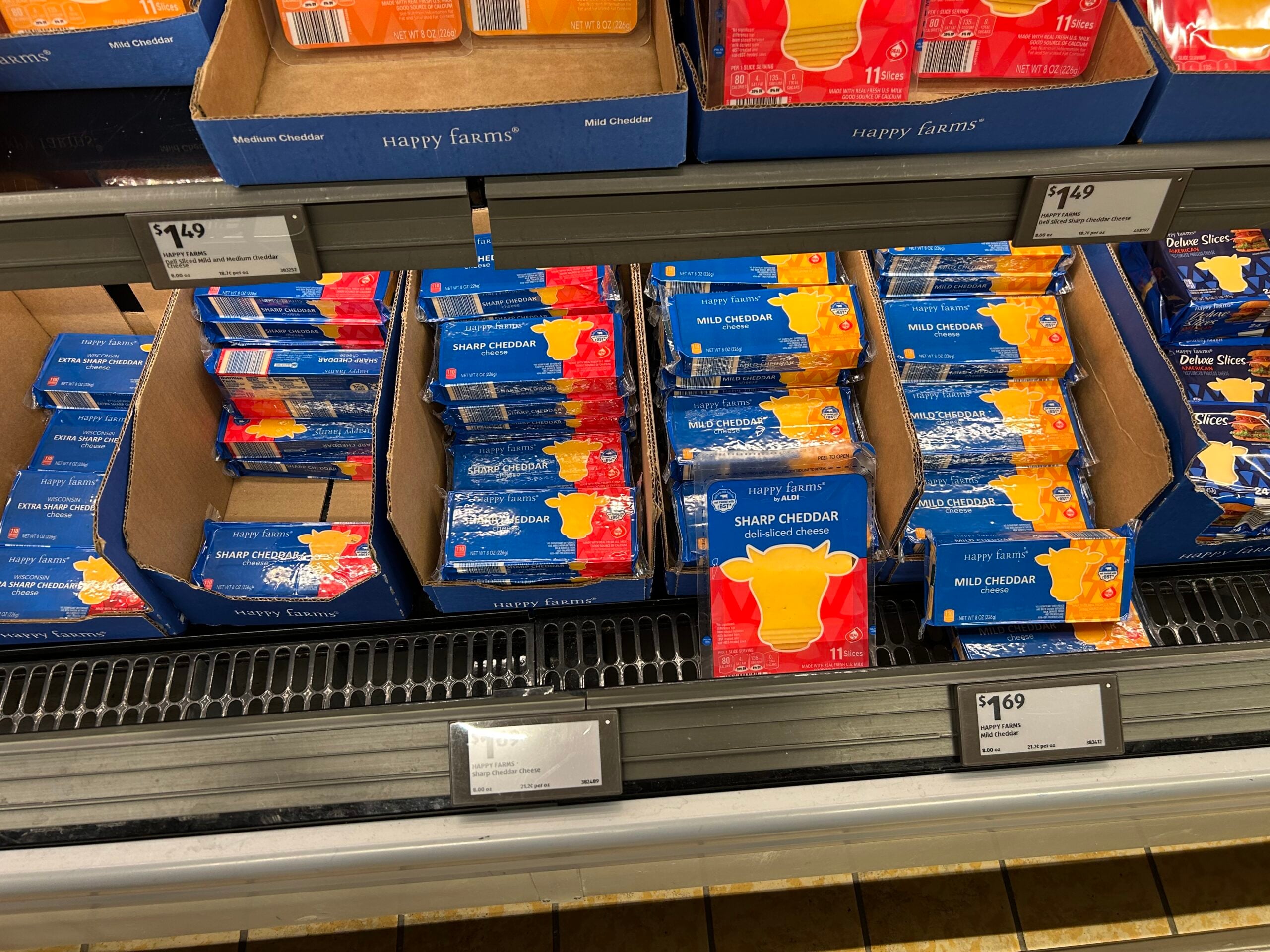 Several shelves display blocks of Happy Farms brand cheese, including sharp cheddar and mild cheddar varieties, packaged in blue and orange wrappers inside cardboard boxes in a refrigerated section. Price tags are visible below.