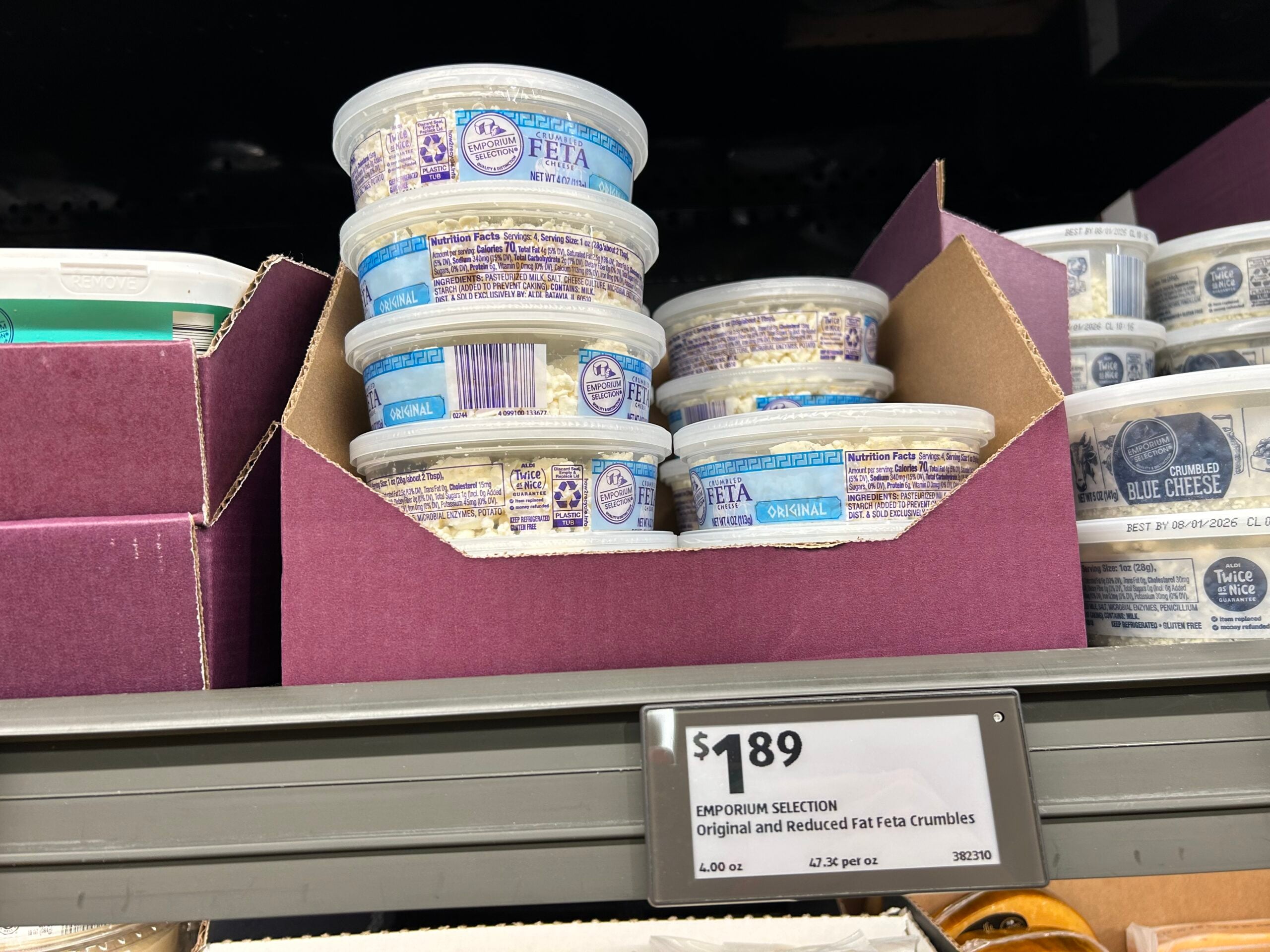 Plastic tubs of Emporium Selection original and reduced fat feta crumbles are stacked in a purple display box on a store shelf. A price tag below reads $1.89 per 4-ounce container.