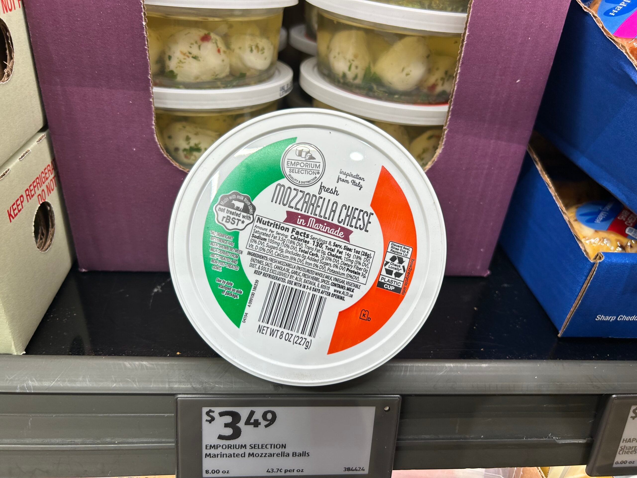 A round container of marinated mozzarella cheese balls with a red, white, and green lid sits on a store shelf. A price tag below reads "$3.49 Emporium Selection Marinated Mozzarella Balls.