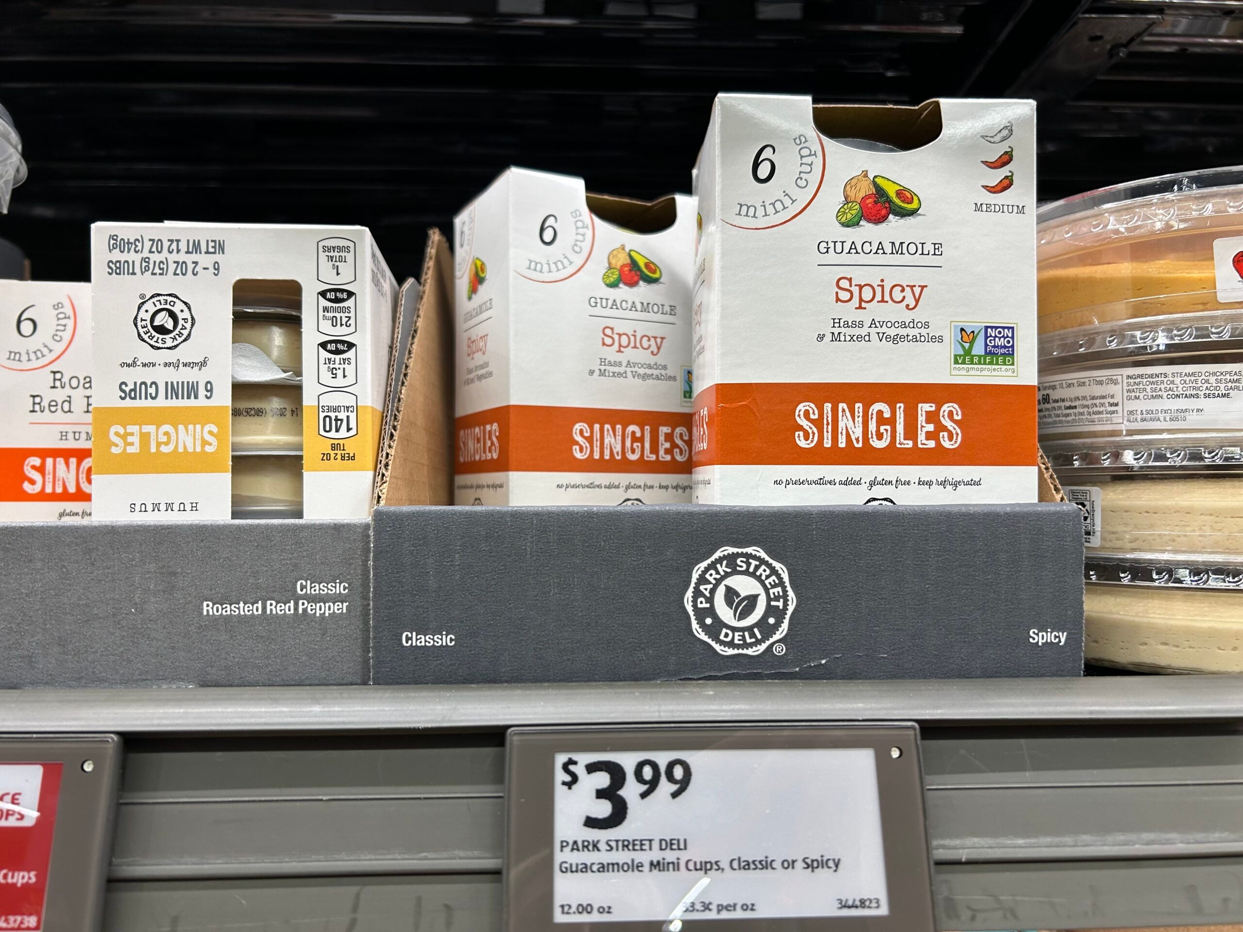 Boxes of Park Street Deli Guacamole Mini Cups in Spicy flavor are displayed on a grocery store shelf with a price tag showing $3.99. The packaging highlights that each box contains six mini cups.