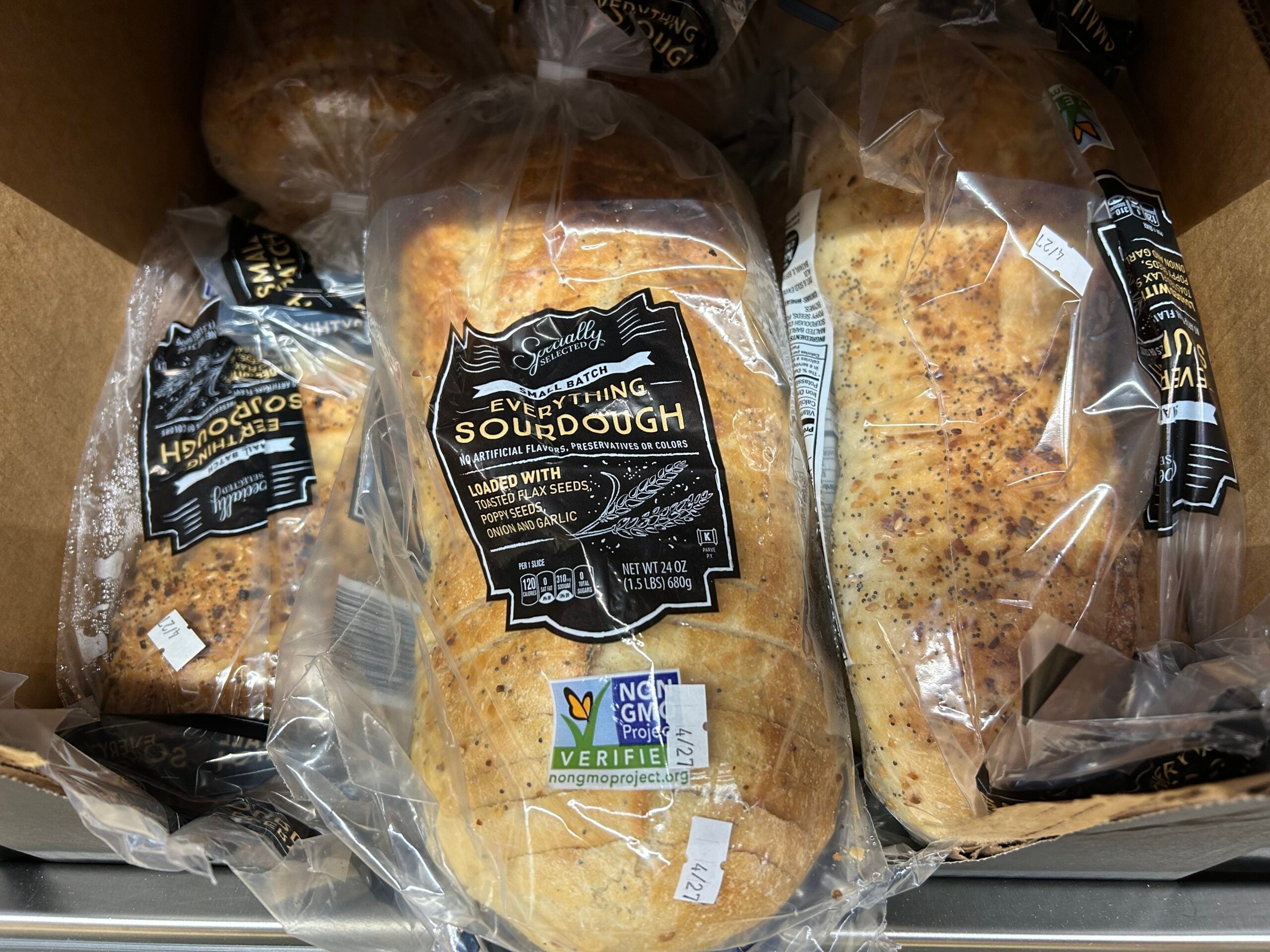 Loaves of Everything Sourdough bread in clear plastic packaging with black and white labels, displayed in a cardboard box. The bread is labeled as non-GMO and features visible seeds and seasonings on the crust.