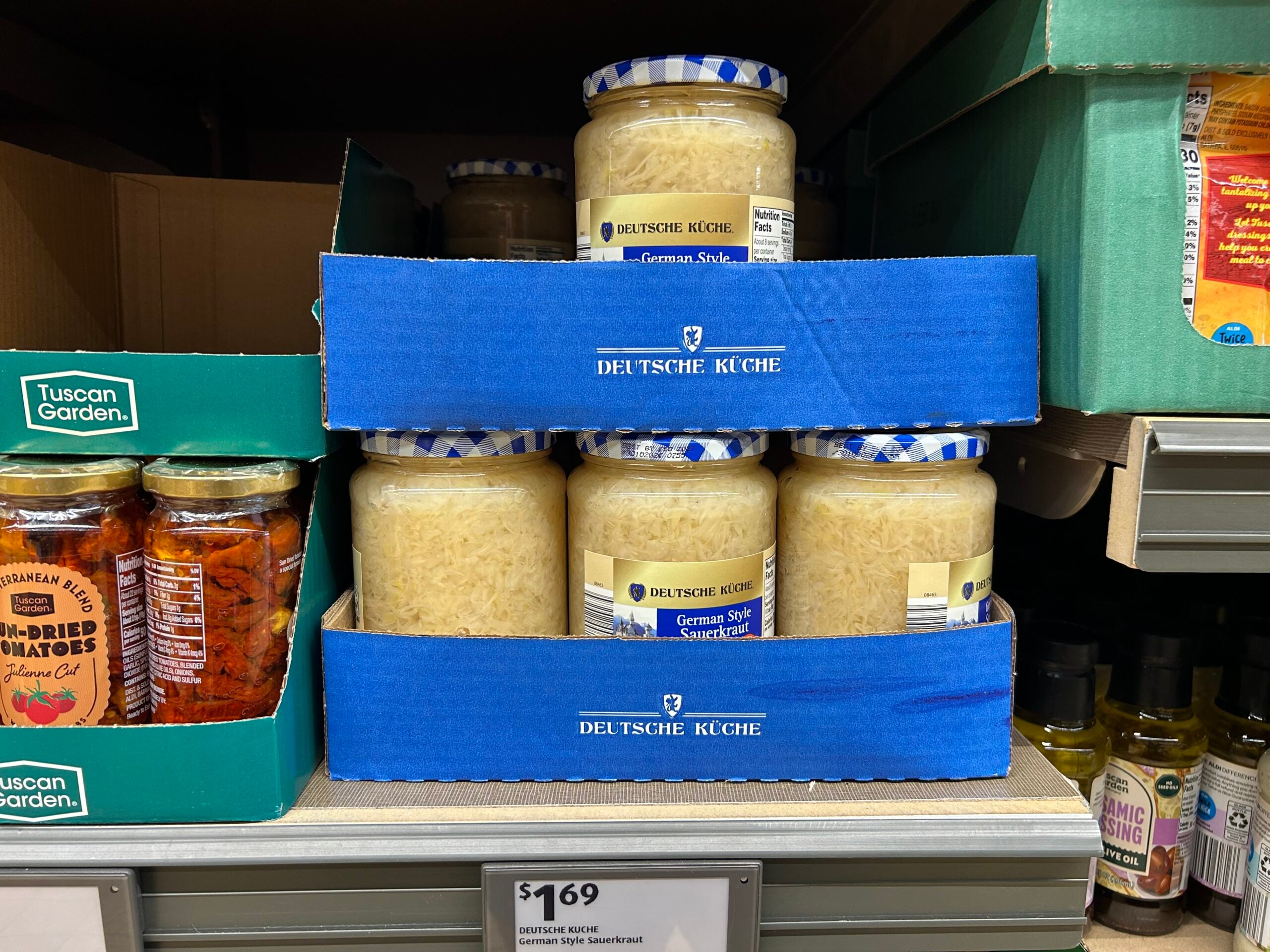 Jars of Deutsche Küche German Style Sauerkraut are stacked on a store shelf in blue cardboard trays, with other jars of sun-dried tomatoes and olive oil nearby. The price label below reads $1.69.