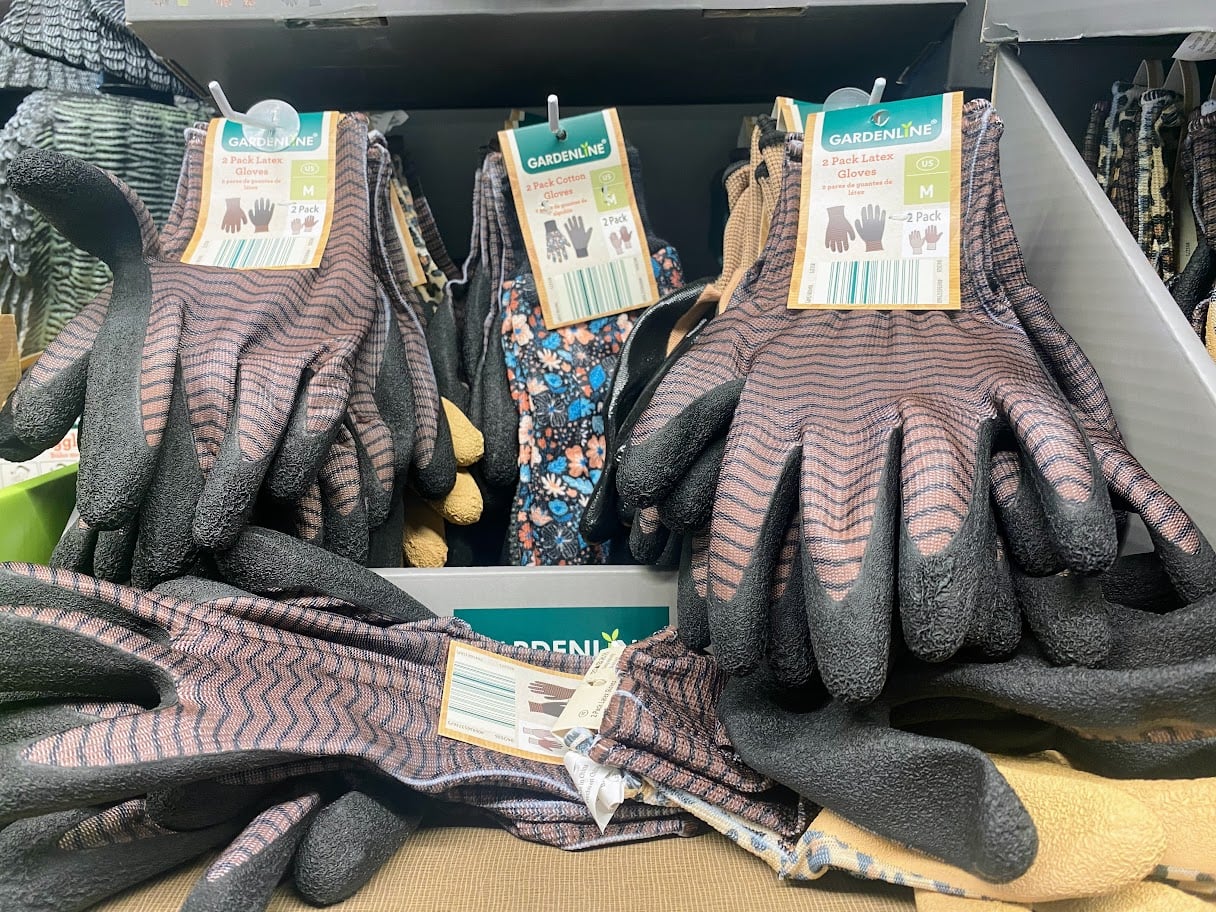 Several pairs of brown and black textured gardening gloves with zigzag patterns are displayed in a store box. The gloves are labeled “Gardenline” and come in packs of four, with one floral patterned pair visible.