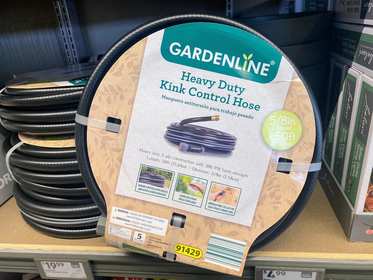 A coiled Gardenline Heavy Duty Kink Control Hose in packaging is displayed on a store shelf. The label highlights it is 5/8 inch wide and 50 feet long, showing features and product information.