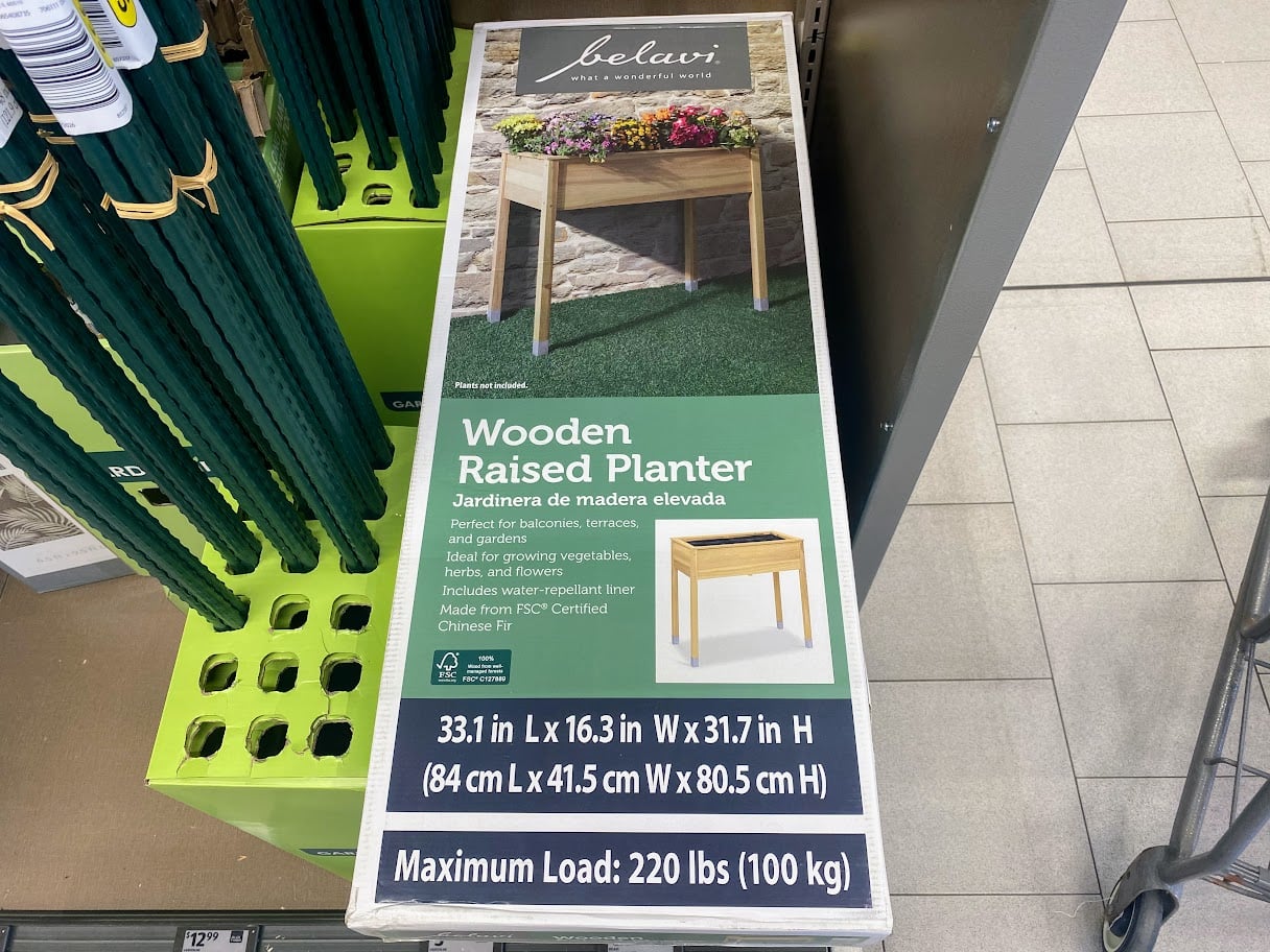 A box for a belavi wooden raised planter on a store shelf, displaying dimensions, features, and a photo of the assembled planter with plants inside. Text indicates a maximum load of 220 lbs (100 kg).