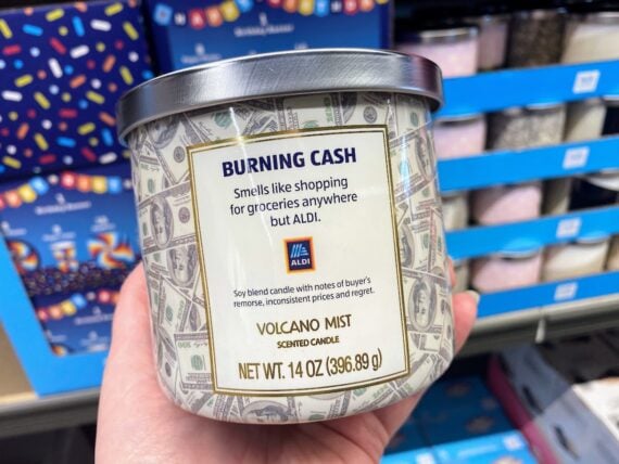 A hand holds a candle labeled "BURNING CASH," which reads, "smells like shopping for groceries anywhere but ALDI." The candle is decorated with dollar bills and is scented "Volcano Mist." Shelves of candles are in the background.