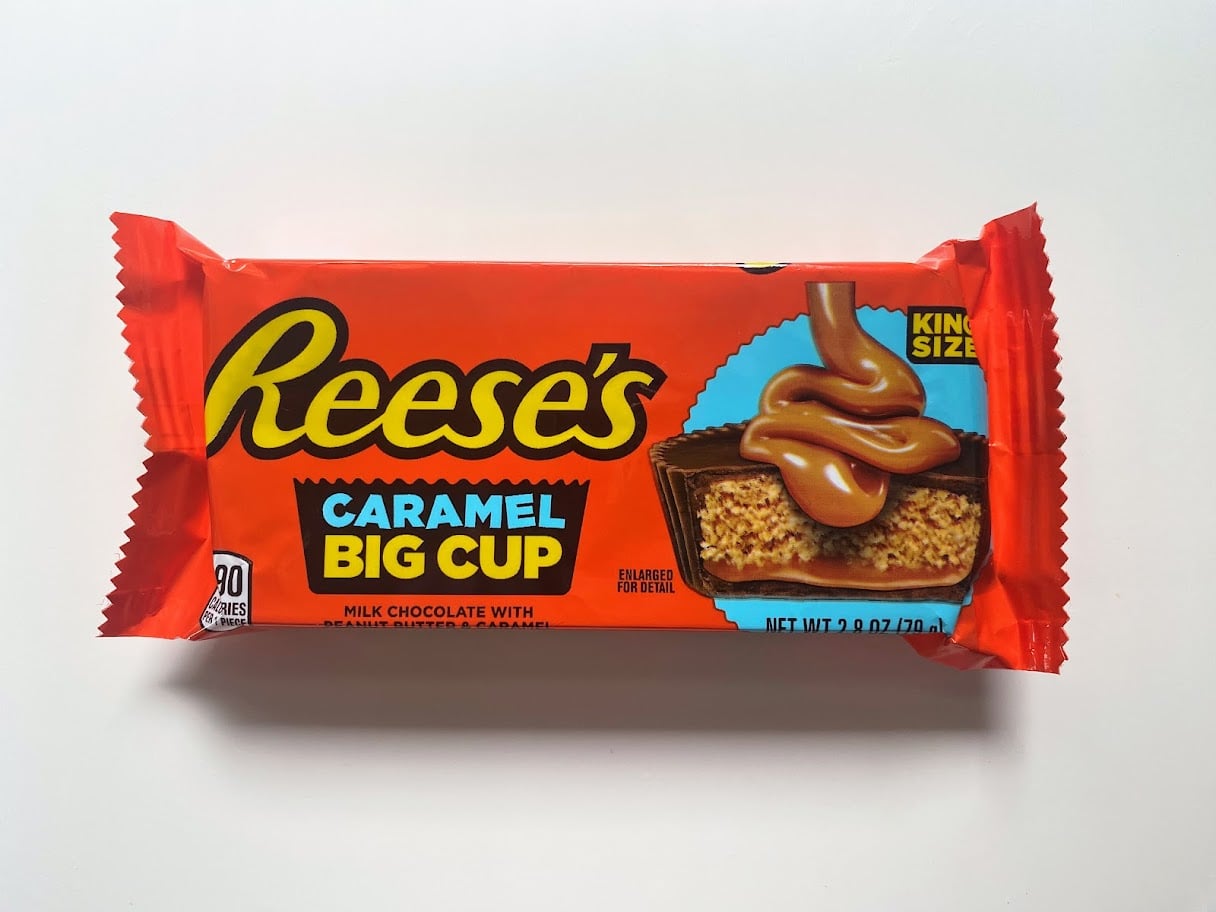 A king size Reese's Caramel Big Cup candy bar in orange packaging, featuring an image of a chocolate cup filled with peanut butter and caramel flowing on top.