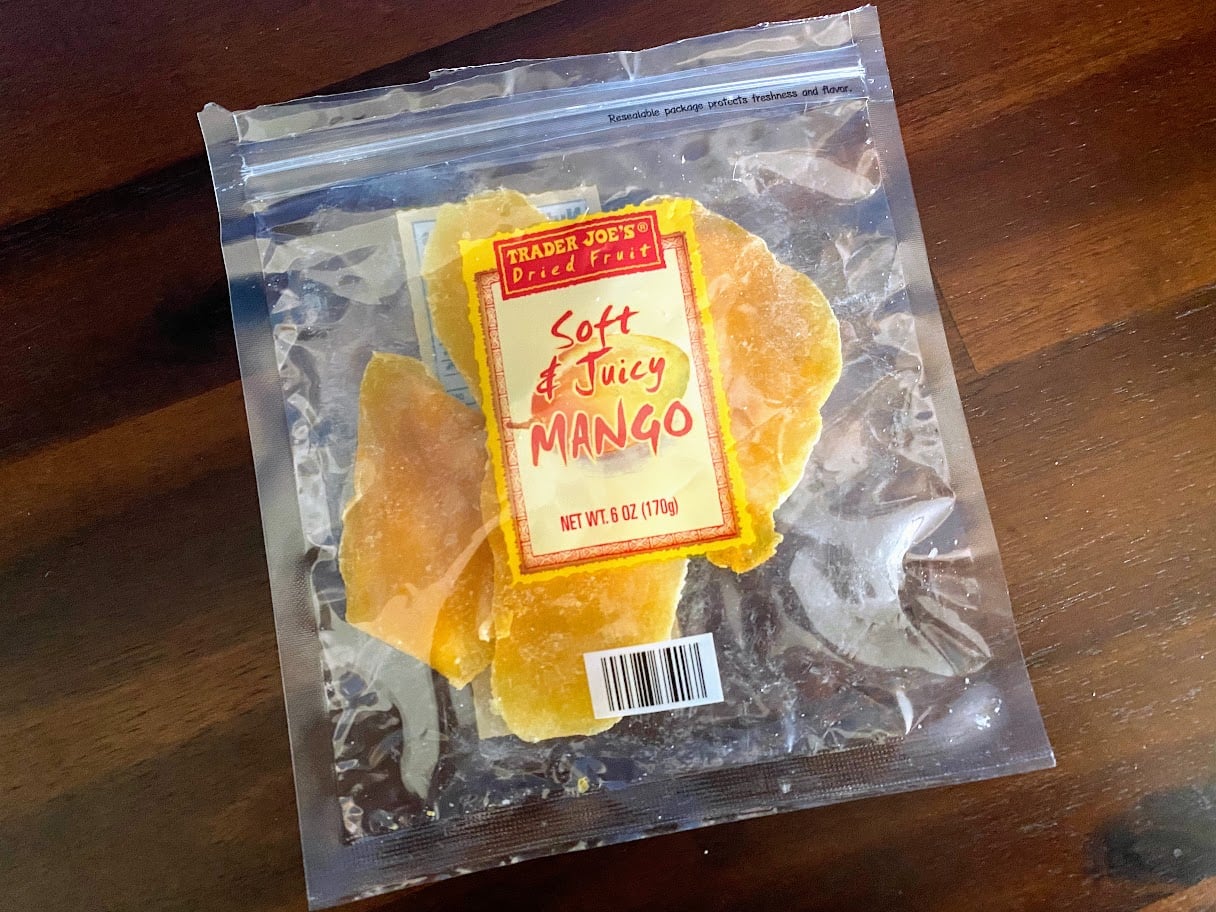 A clear resealable bag of Trader Joe’s Soft & Juicy Mango dried fruit sits on a dark wooden surface. The bag is partially filled with bright orange mango slices visible through the packaging.