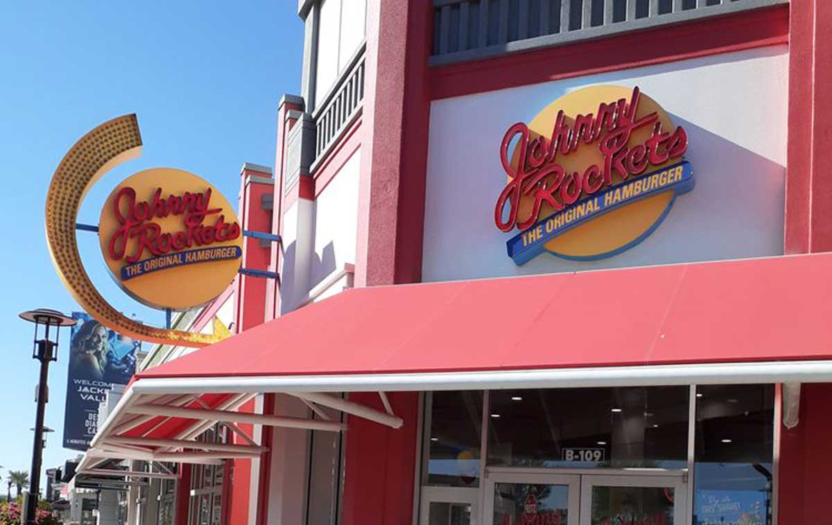 The exterior of a Johnny Rockets restaurant, reminiscent of old-school diners in America, features a large yellow and red sign reading “Johnny Rockets The Original Hamburger” above the entrance and on a pole under a red awning.