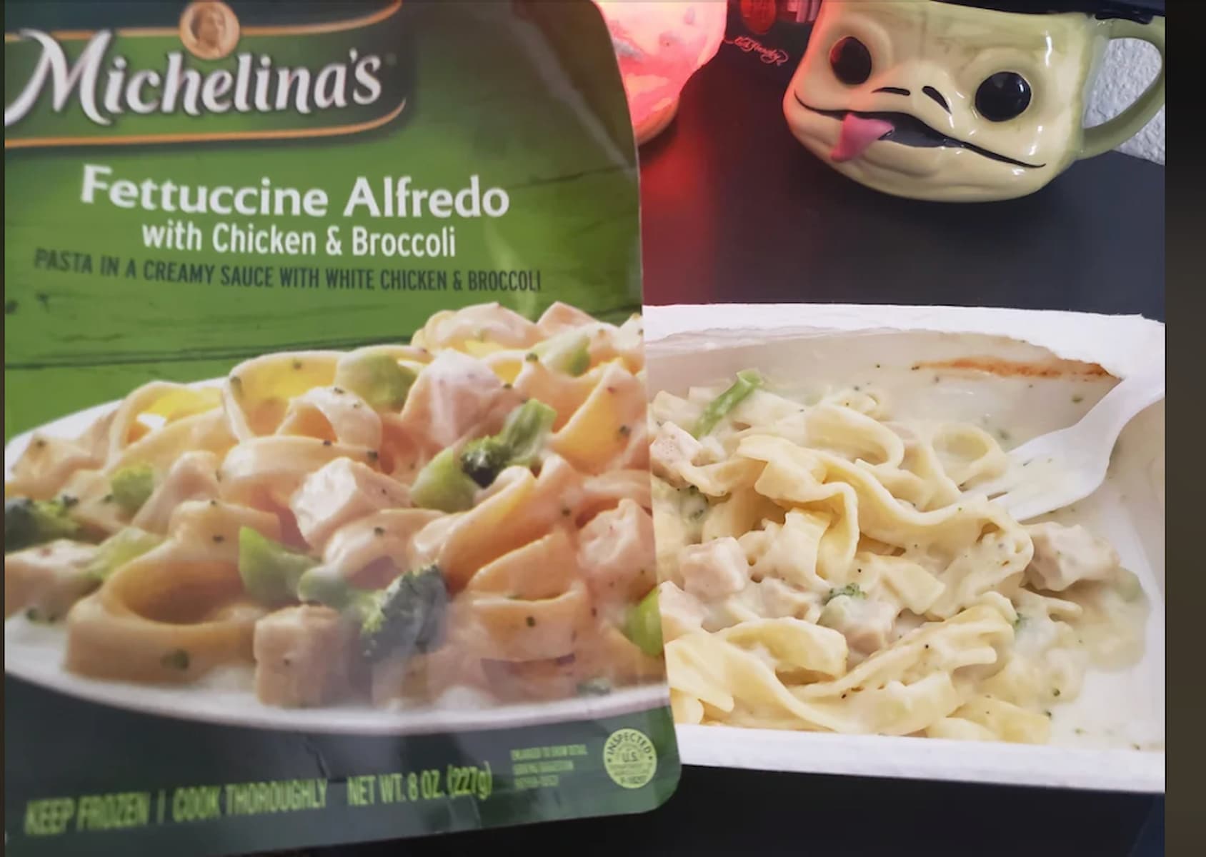 A Michelina’s Fettuccine Alfredo with Chicken & Broccoli frozen meal package is shown beside the cooked meal in a tray. A mug with a cartoon animal face and a lit candle are in the background.