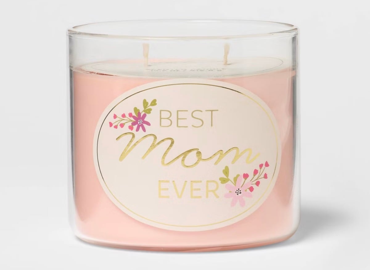 A pink candle in a clear glass jar with a label that reads "Best Mom Ever" in gold and pink, decorated with small pink flowers and green leaves—perfect for mother’s day gifts.