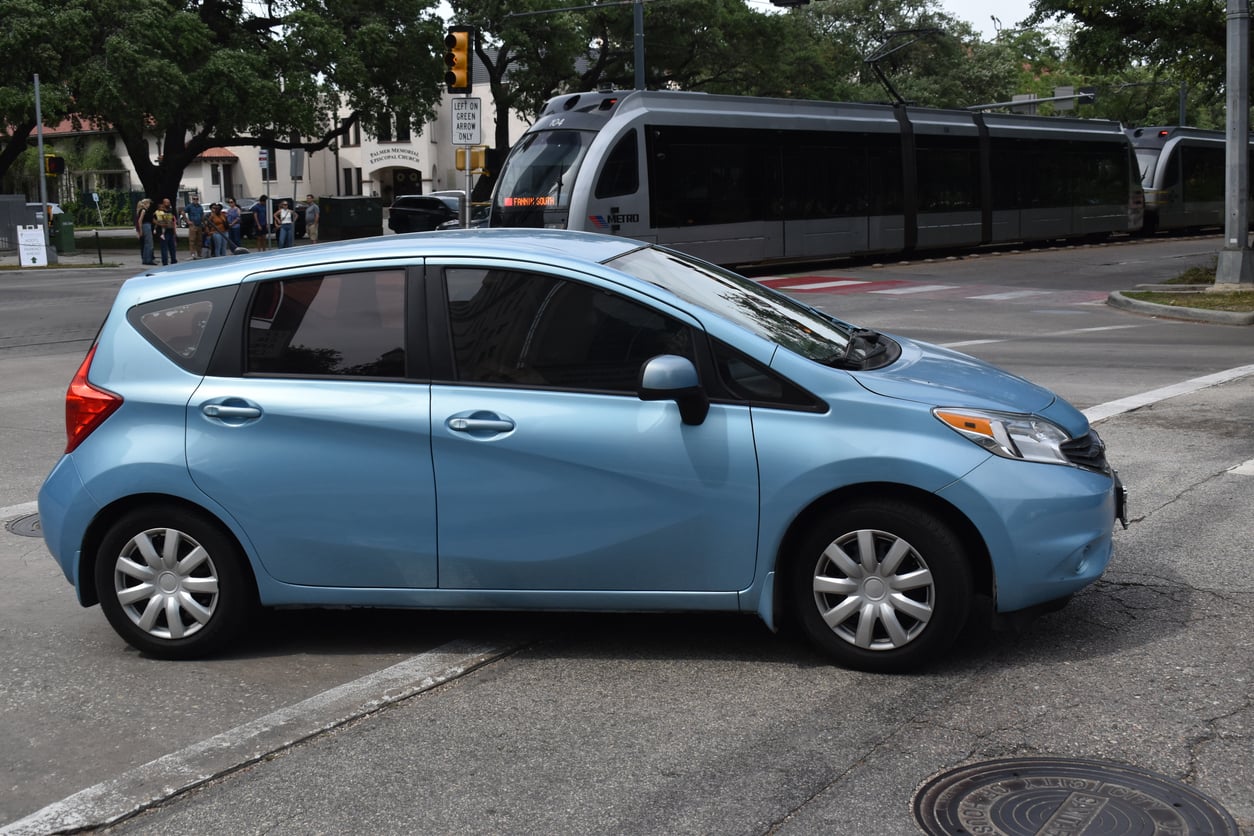 A light blue compact car, one of the new cars on the road, waits at an intersection, partially blocking a crosswalk. In the background, a light rail train is stopped on the tracks, with trees and buildings visible.