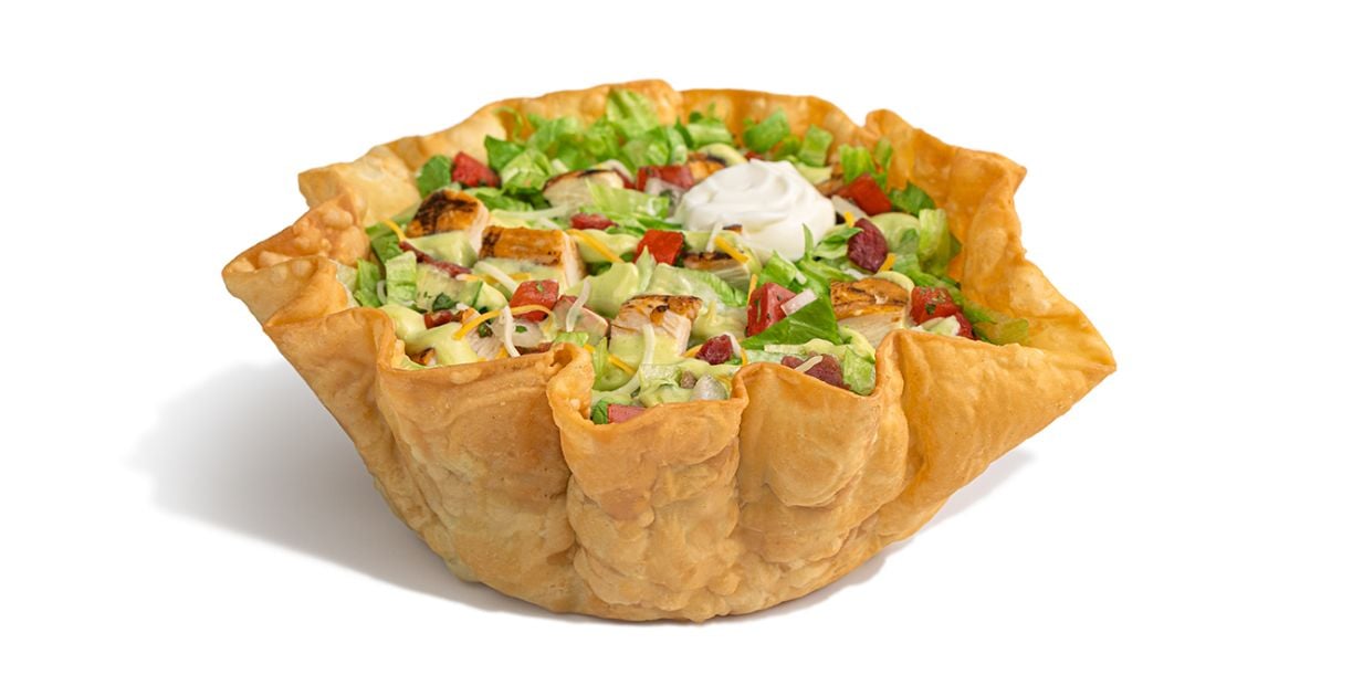A crispy tortilla bowl from our new fast-food menu, filled with lettuce, grilled chicken pieces, diced tomatoes, cheese, and topped with a dollop of sour cream, isolated on a white background.