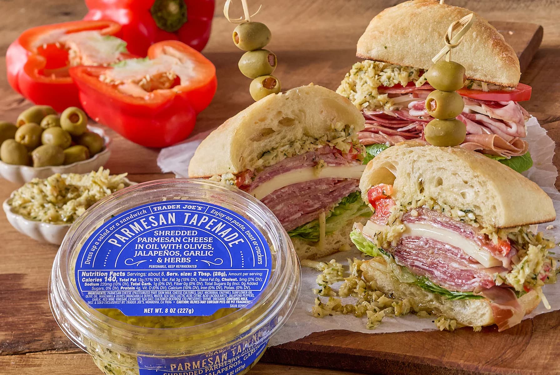 A container of Parmesan tapenade sits next to Italian sandwiches filled with meats, cheese, and lettuce on ciabatta rolls, garnished with green olives. Bowls of tapenade and sliced red bell peppers are in the background.