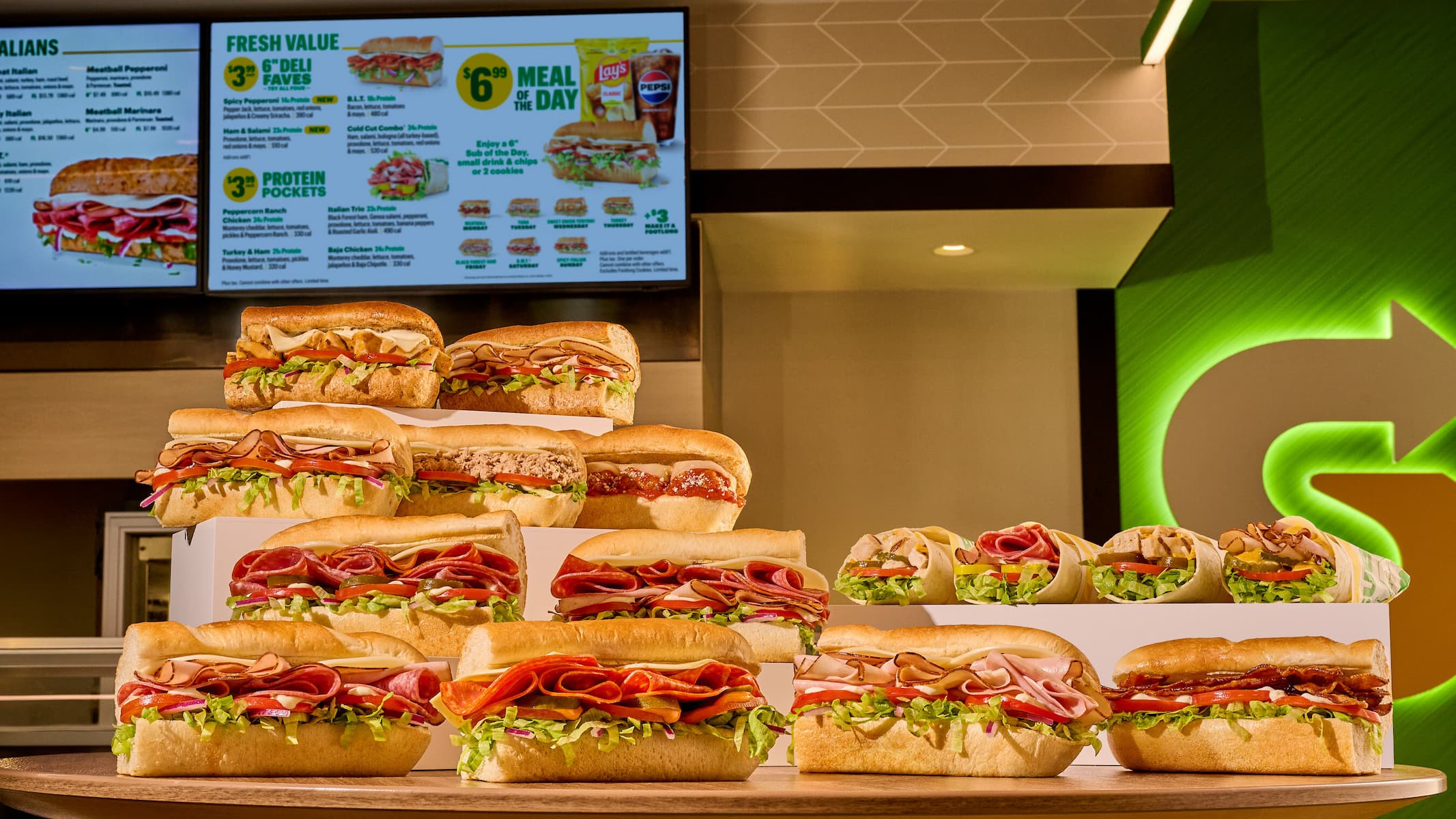 A display of assorted submarine sandwiches with various meats, cheeses, and vegetables is arranged in front of a Subway value menu board inside a brightly lit restaurant.