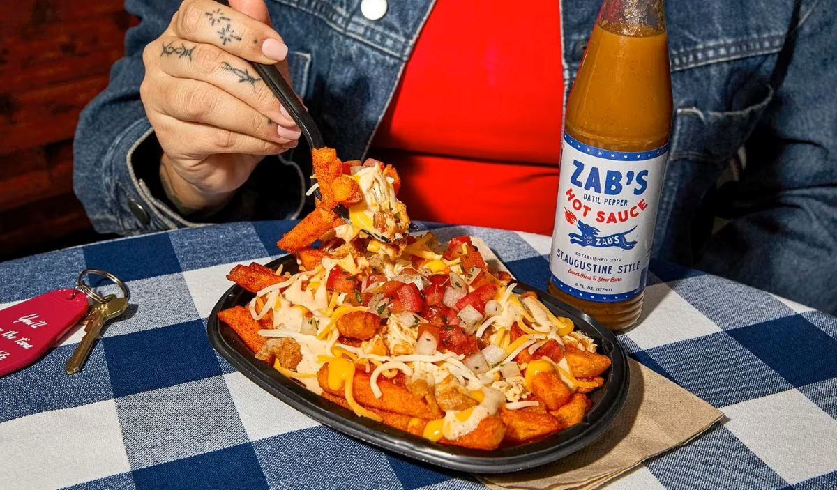 A person in a denim jacket holds a fork with loaded fries topped with cheese, tomatoes, and meat from the new fast-food menu. A bottle of Zab's Hot Sauce sits nearby on a blue-and-white checkered tablecloth.
