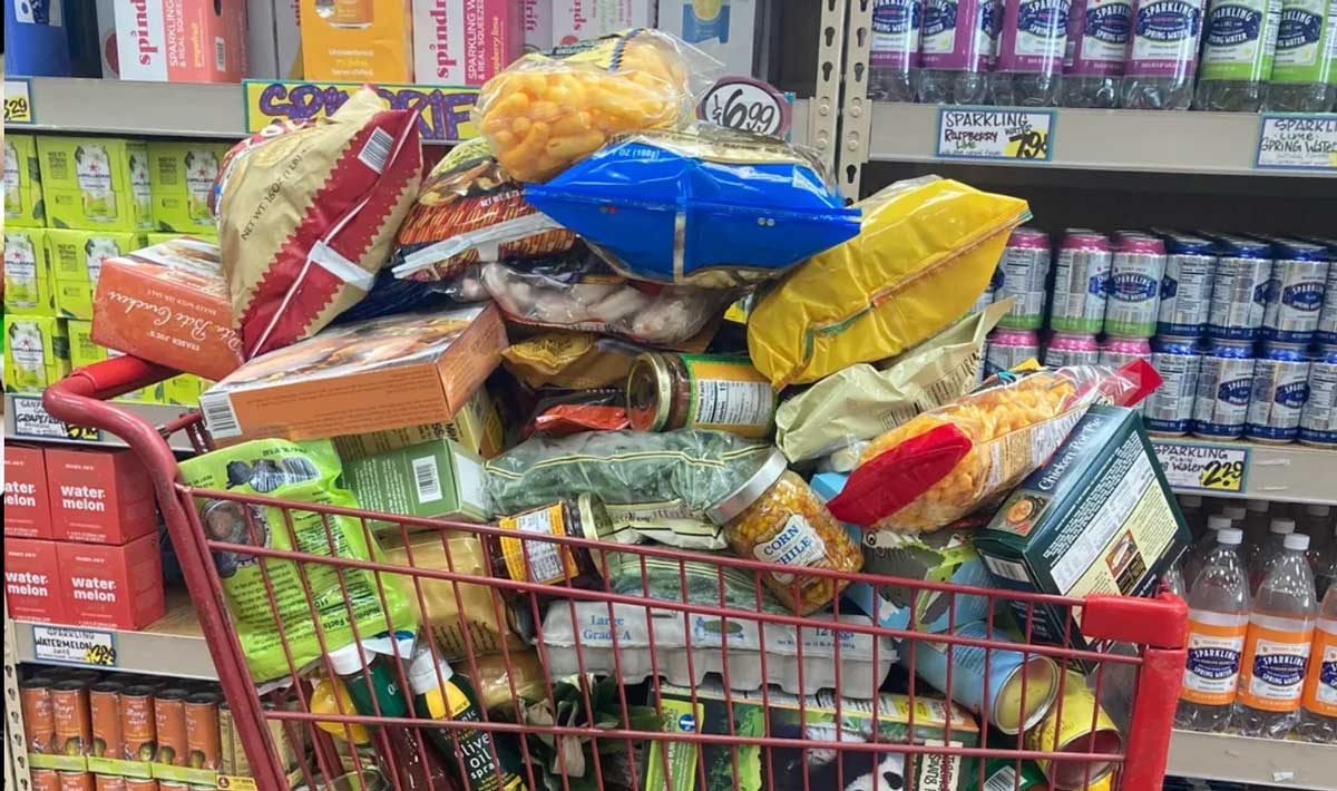 A grocery cart overflowing with Trader Joe's groceries—various packaged foods, canned goods, and snacks—sits in a supermarket aisle lined with beverages and other grocery items.