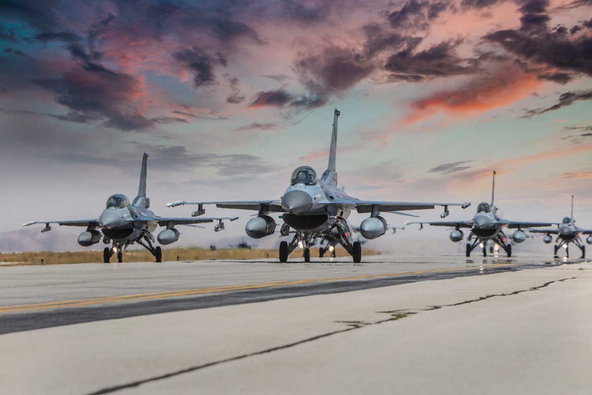 Four fighter jets taxi on a runway under a dramatic sky at sunset, with clouds displaying shades of pink, orange, and purple. The jets are lined up, facing forward, ready for takeoff.
