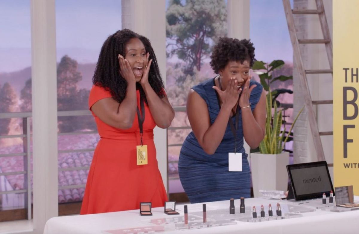 Two women stand behind a table displaying makeup products, looking excited with their hands on their faces. Wearing dresses and event badges, they showcase beauty items and discuss staying stylish even after overcoming bankruptcy.