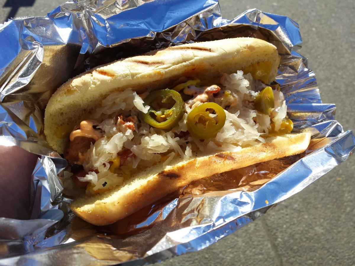 A grilled hot dog in a bun, topped with sauerkraut, sliced jalapeños, and sauce, served on a sheet of crinkled aluminum foil.
