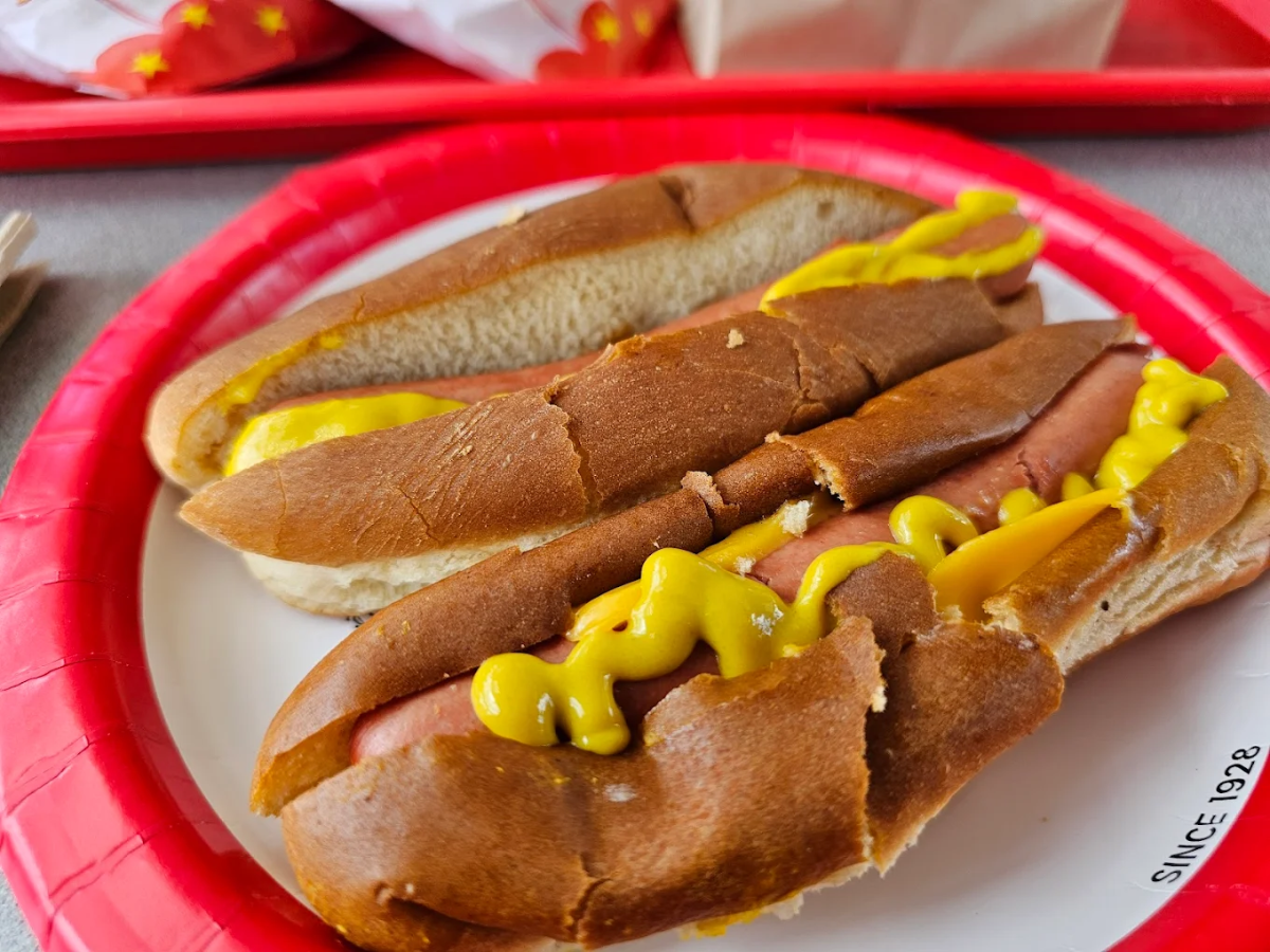 Two hot dogs in split buns topped with yellow mustard, served on a red and white paper plate. The buns appear slightly cracked and unevenly cut.